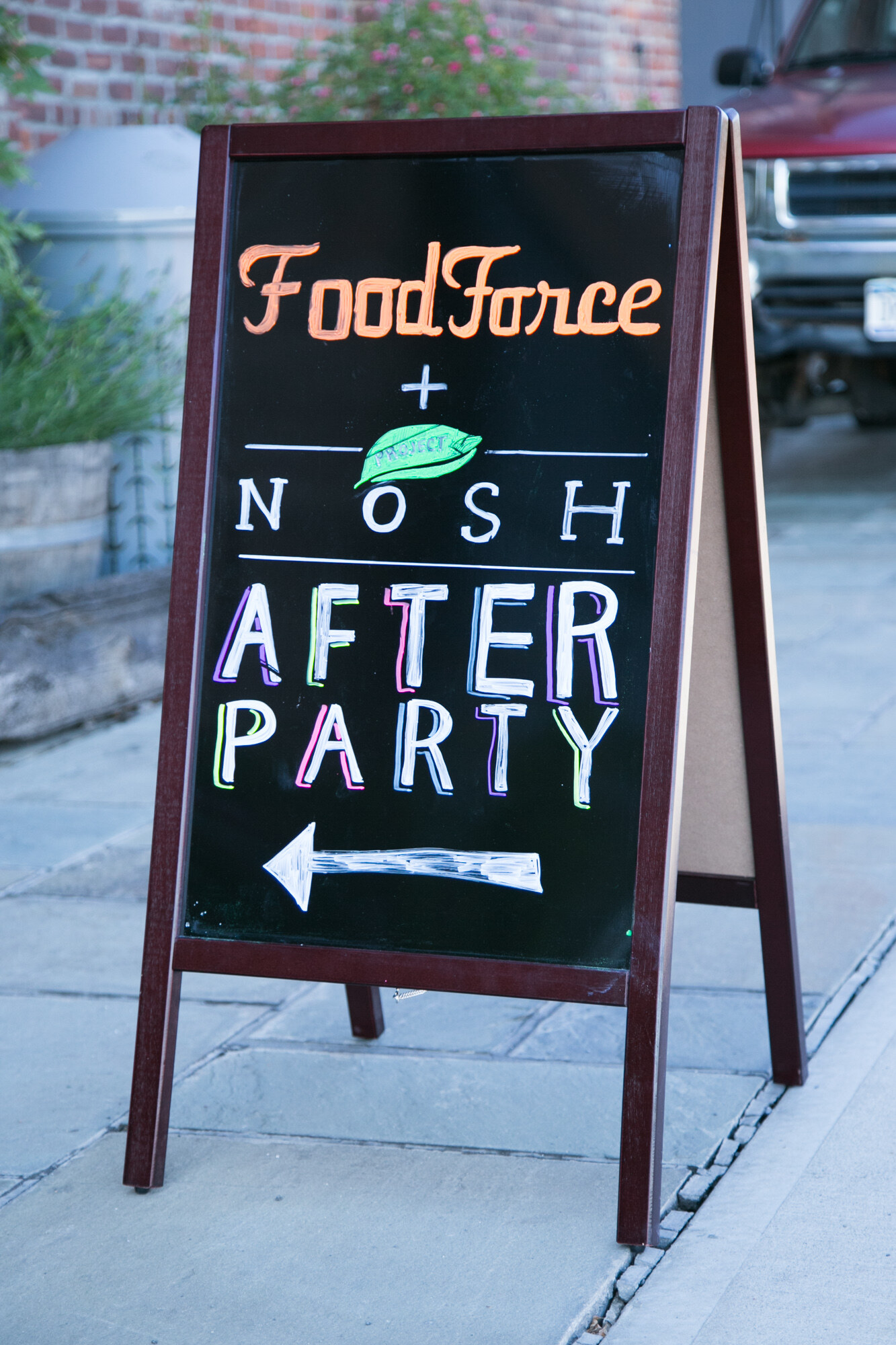 Chalkboard sandwich board reading Food Force plus NOSH After Party with an arrow, on a sidewalk.