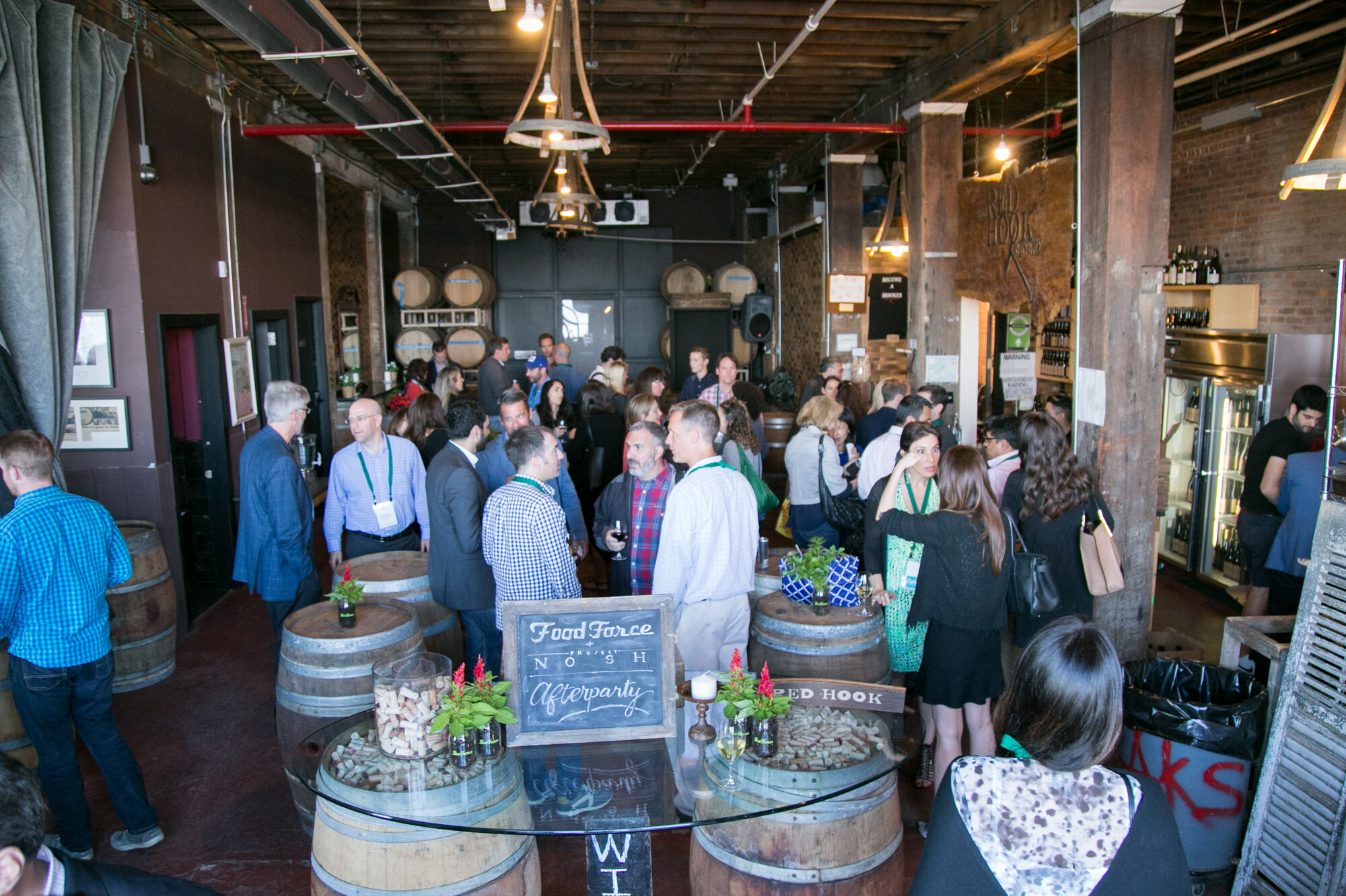 Wide view of guests standing around wine barrels in a brick room with a Food Force Project NOSH Afterparty chalkboard sign on a table.