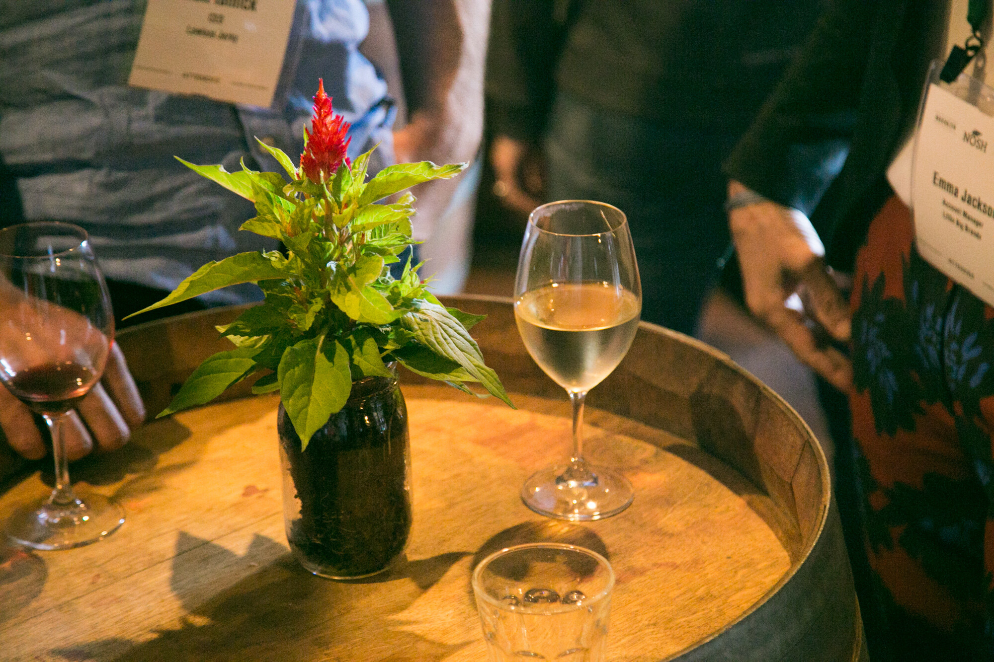 Barrel table with a wine glass, a red flowering plant in a jar and attendee name badges around it.