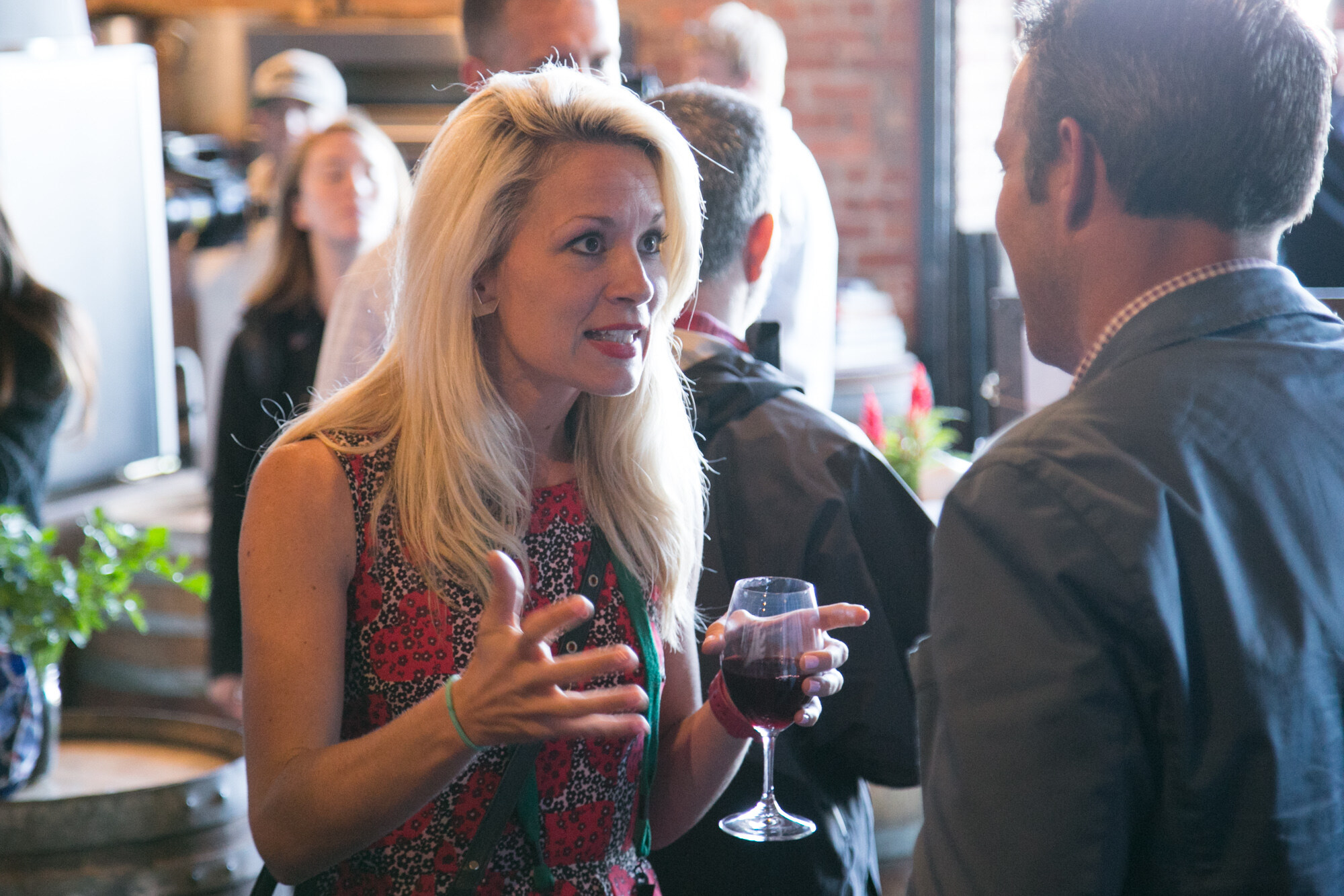 Blonde woman in a red patterned dress holding a wine glass and speaking with a man in a gray shirt.