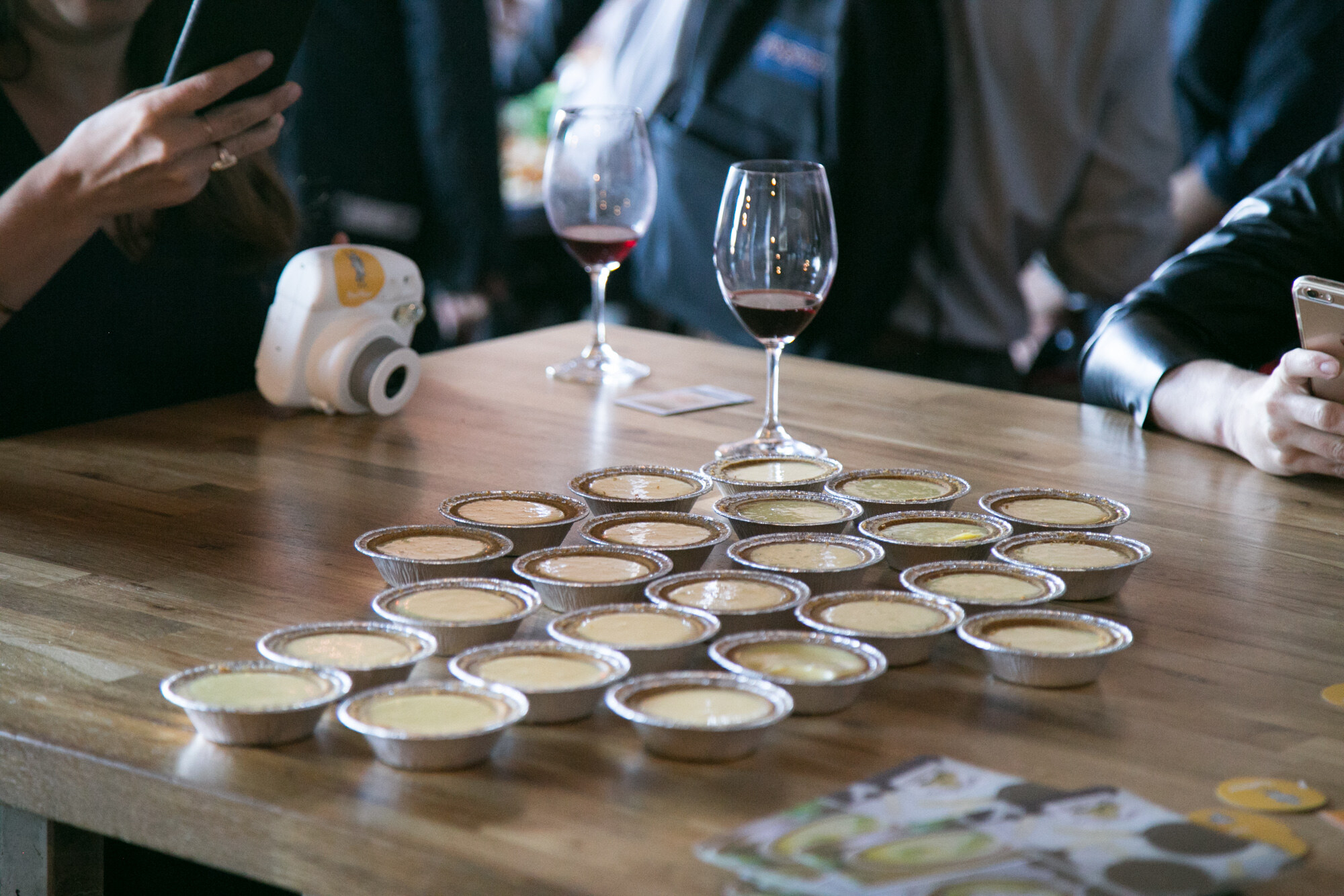 Small foil tasting cups arranged on a wooden table with two glasses of red wine and an instant camera.