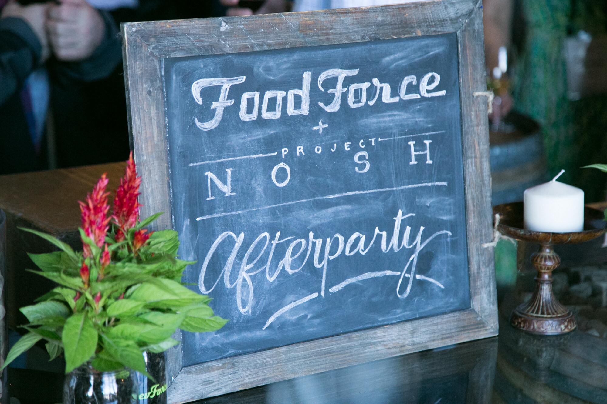 Chalkboard sign reading Food Force plus Project NOSH Afterparty beside a red flowering plant in a jar.
