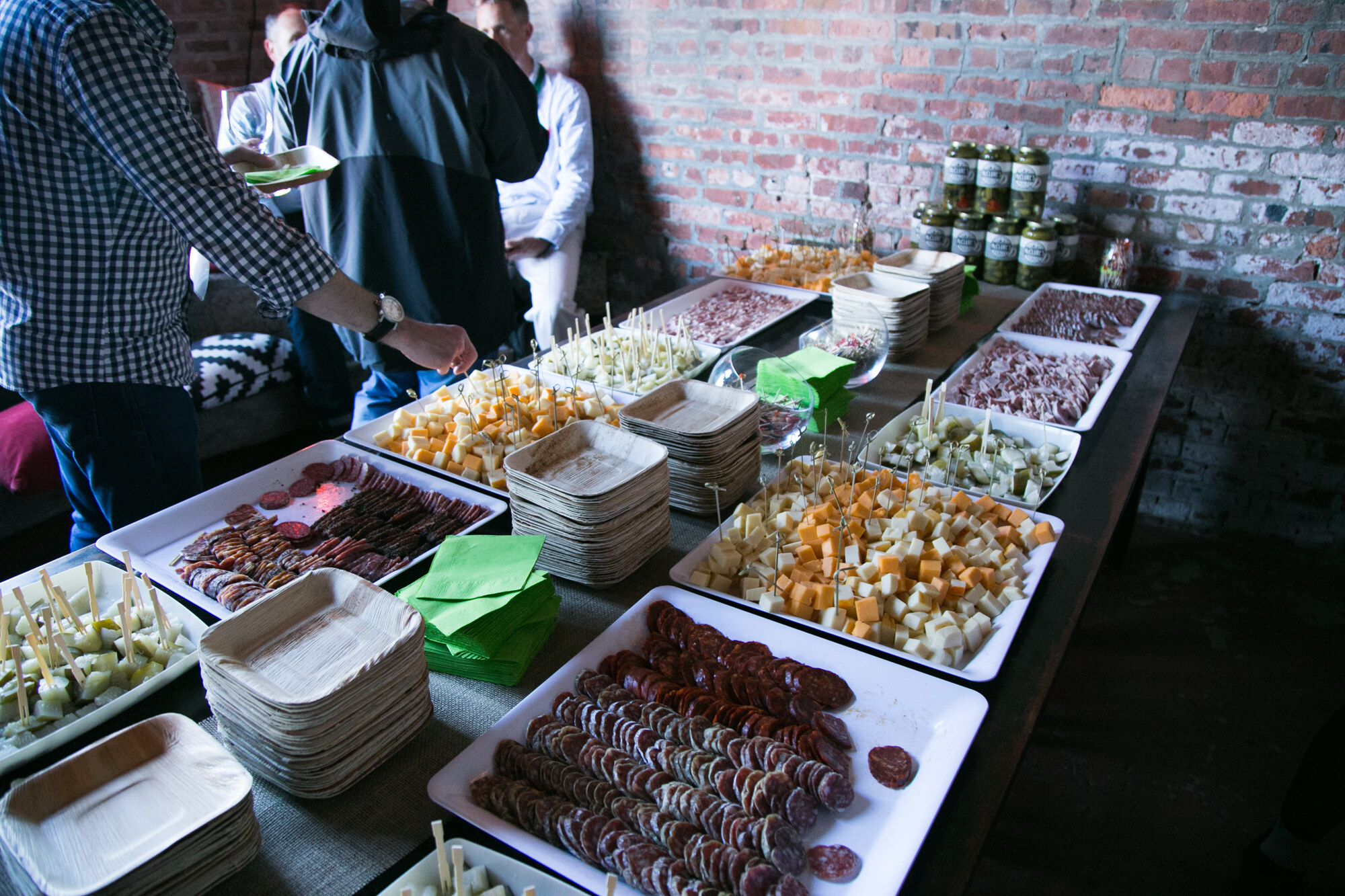 Long table with trays of sliced salami, cheese cubes, cucumber pieces and jars of pickles.