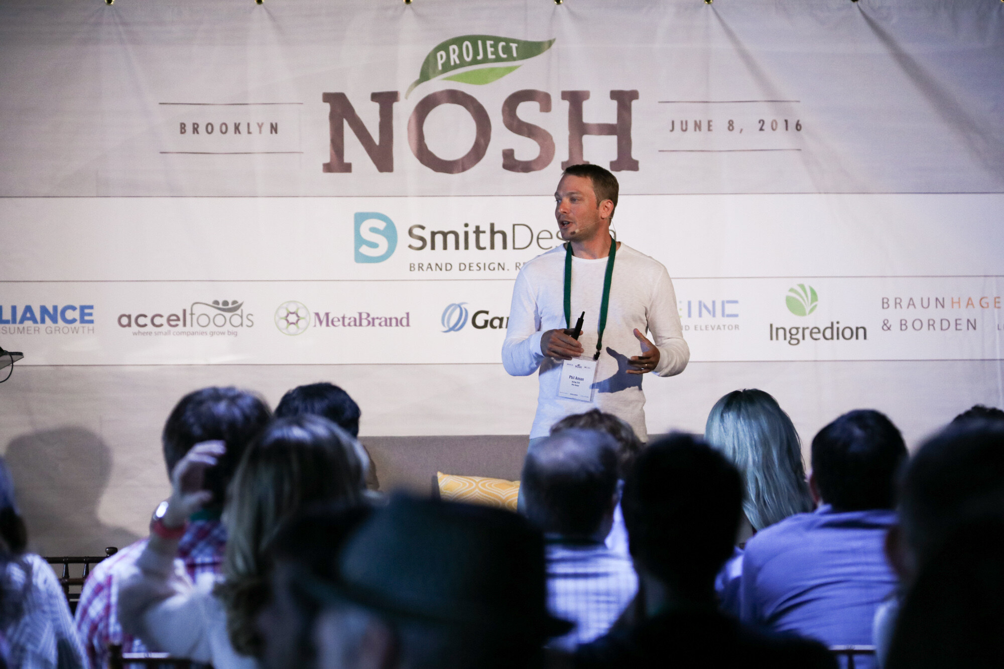 Man in a white long-sleeved shirt and green lanyard speaking on stage under a Project NOSH Brooklyn June 8, 2016 banner, with the backs of audience heads in the foreground.