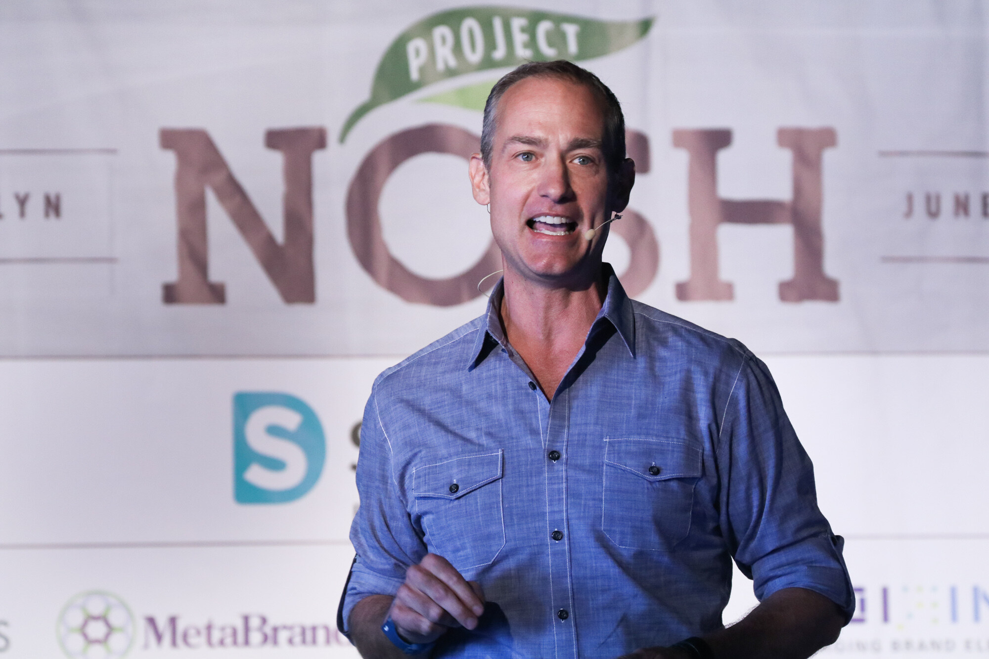 Close view of a man in a blue button-up shirt and headset microphone speaking in front of a large Project NOSH logo.