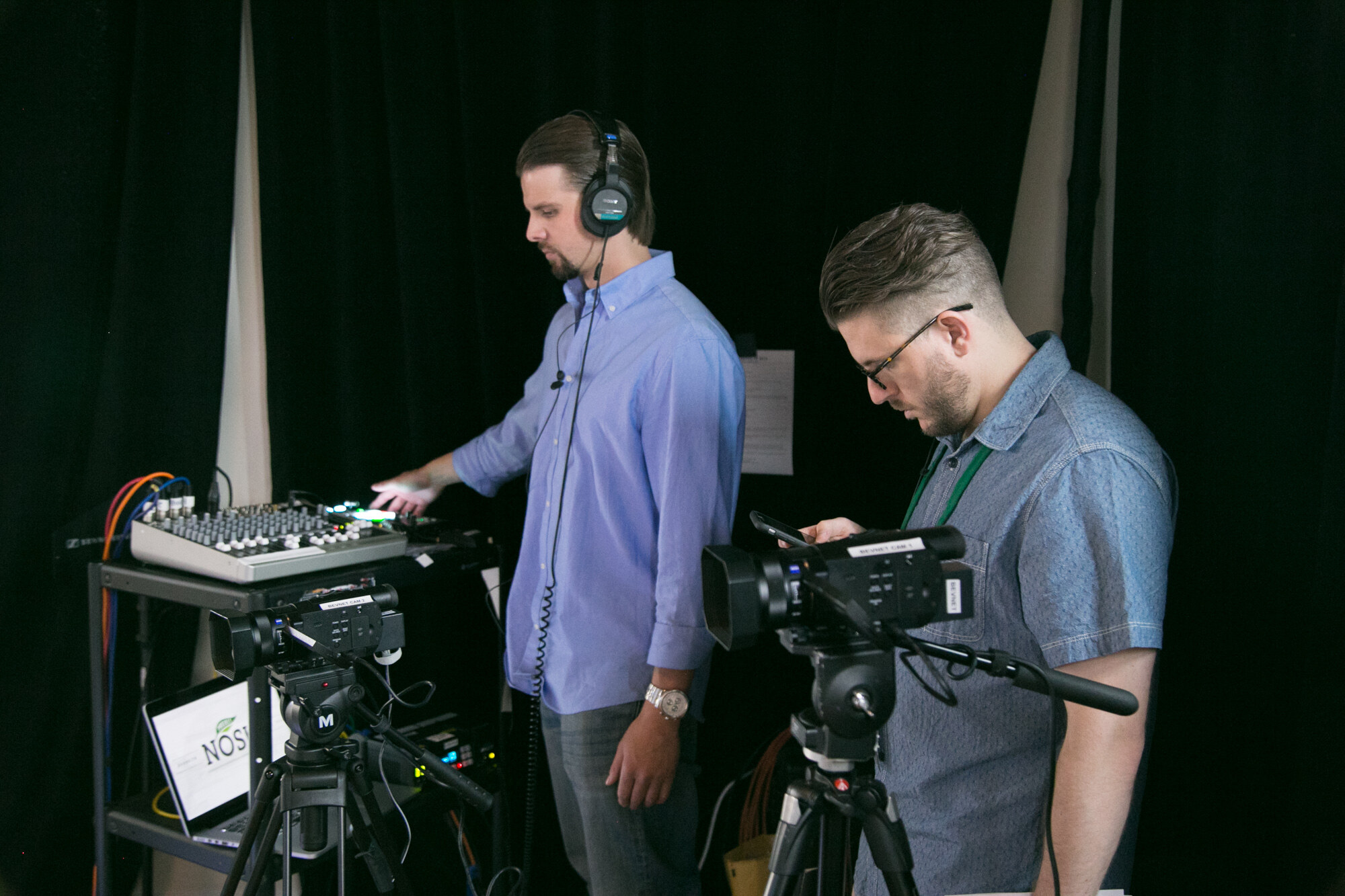 Man in headphones adjusting an audio mixer beside a man in glasses operating a video camera on a tripod, with a laptop showing the NOSH logo.