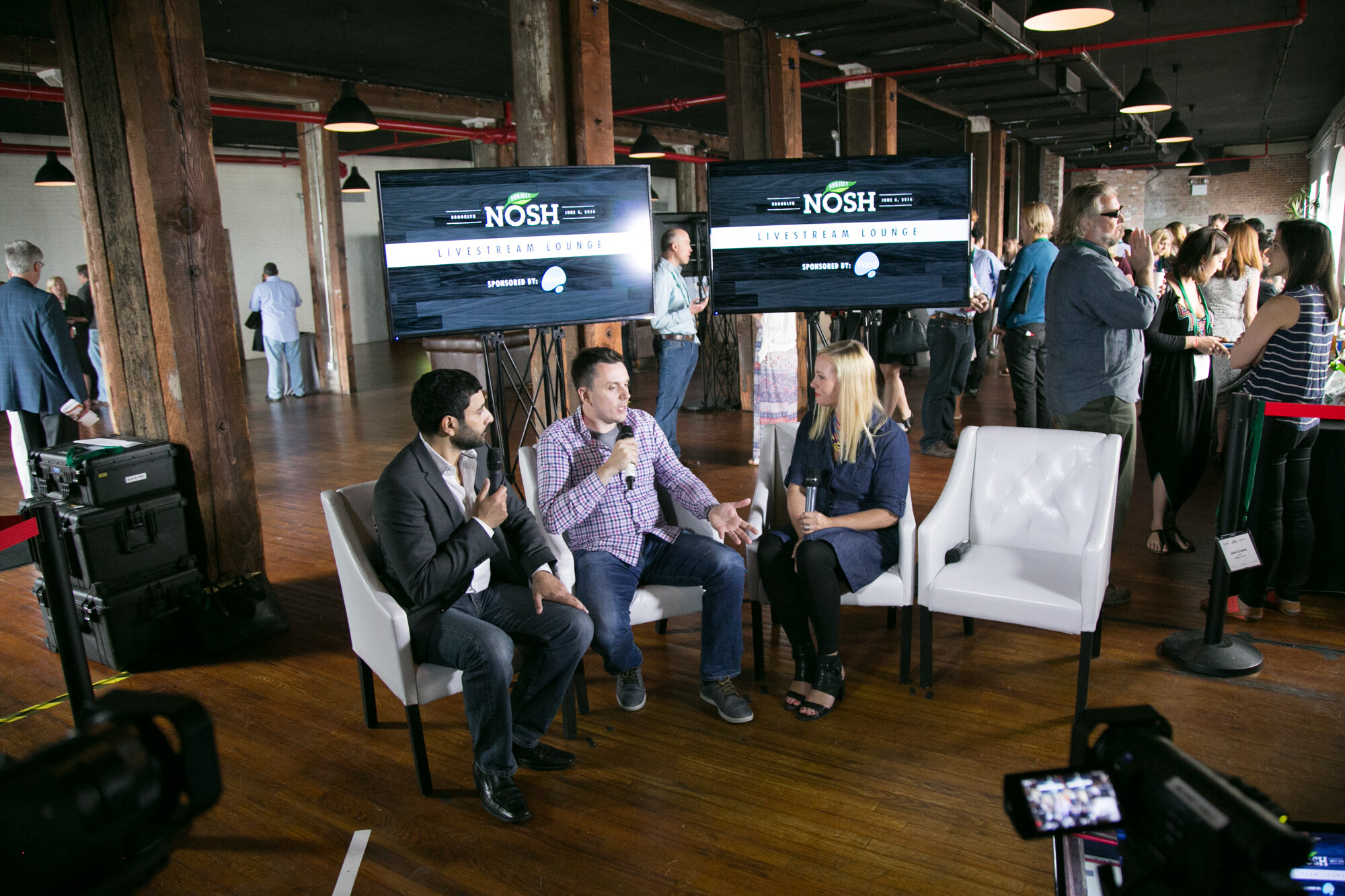 Wide view of the Livestream Lounge with two NOSH screens, three people seated in white chairs holding microphones, and attendees standing behind them.