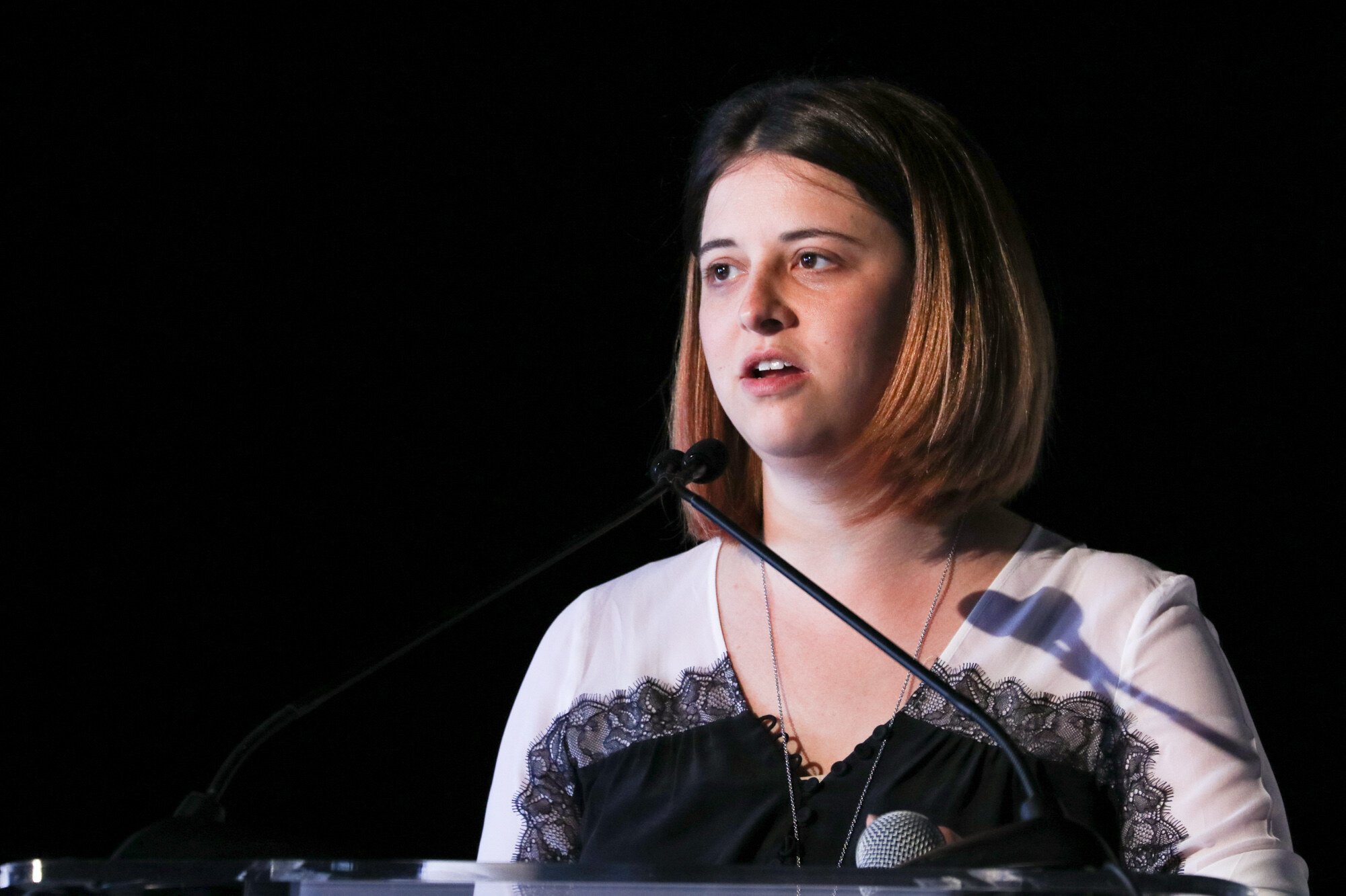 Woman with a bob haircut in a white and black lace top speaking at a clear podium with two microphones.