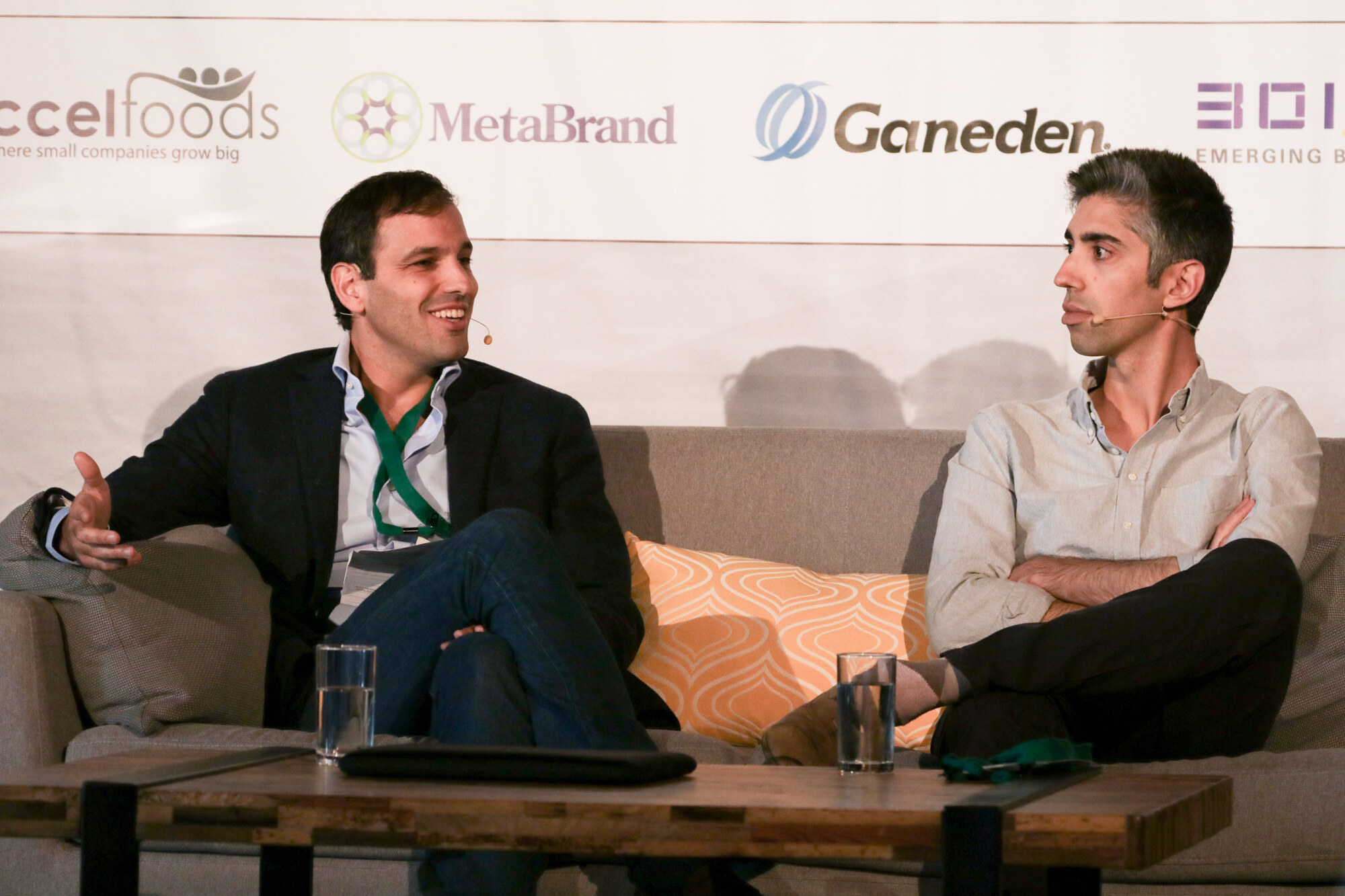 Two men seated on a gray sofa with an orange cushion, one gesturing and one with arms crossed, in front of accelFoods, MetaBrand and Ganeden logos.