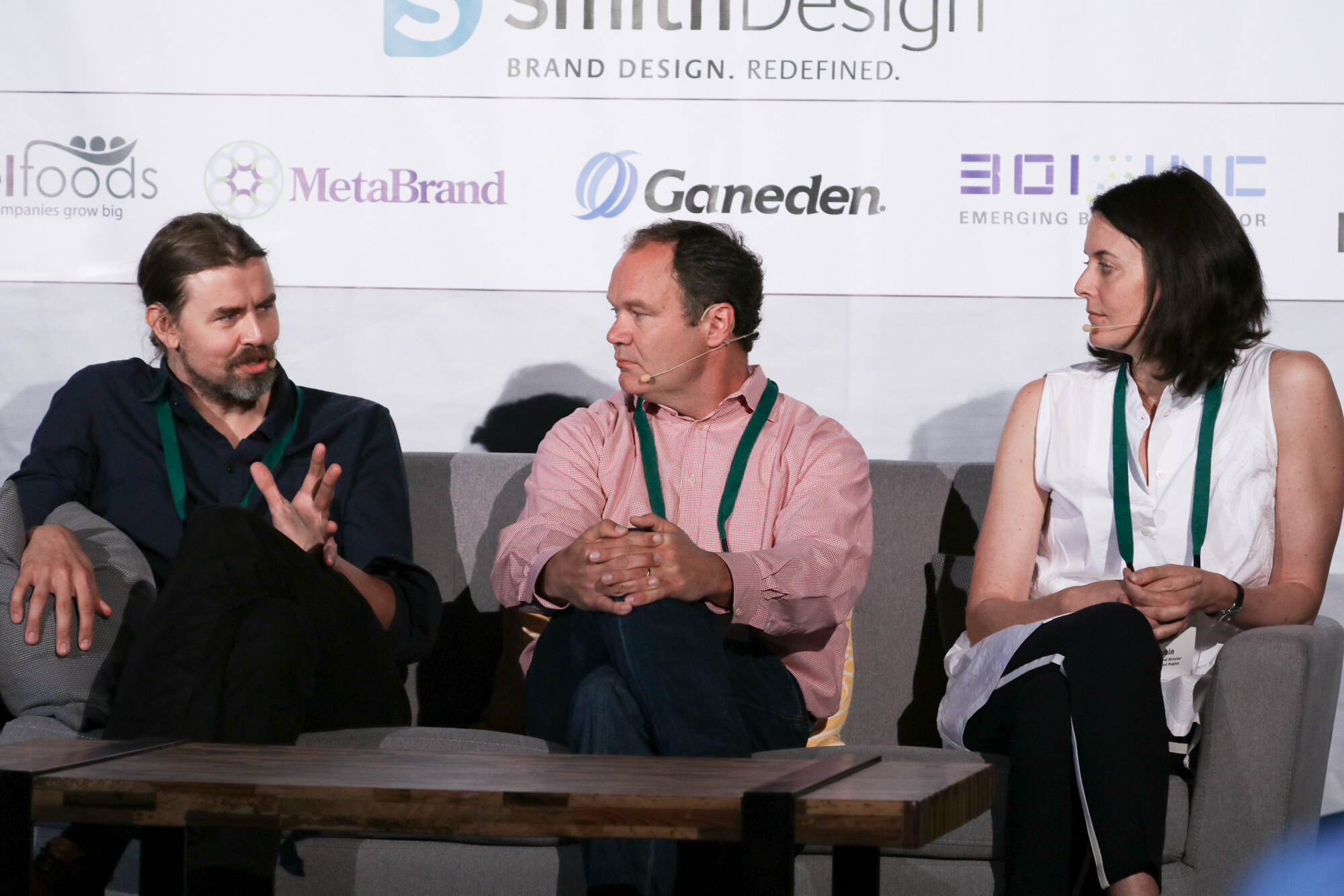 Three people on a gray sofa wearing green lanyards and headset microphones, in front of SmithDesign, MetaBrand and Ganeden logos.