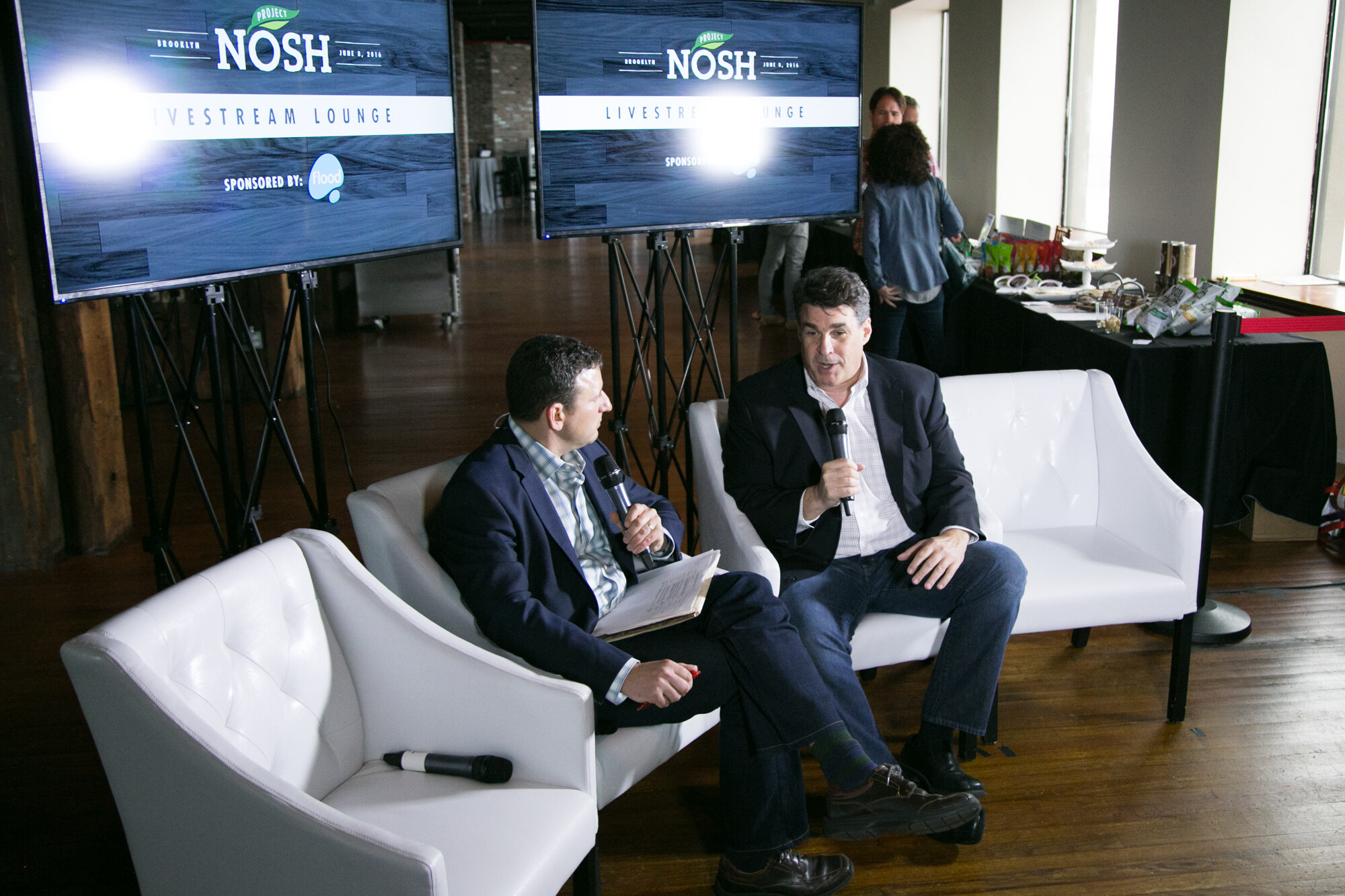 Two men in blazers seated on white chairs with microphones in front of two screens reading Project NOSH Brooklyn Livestream Lounge.