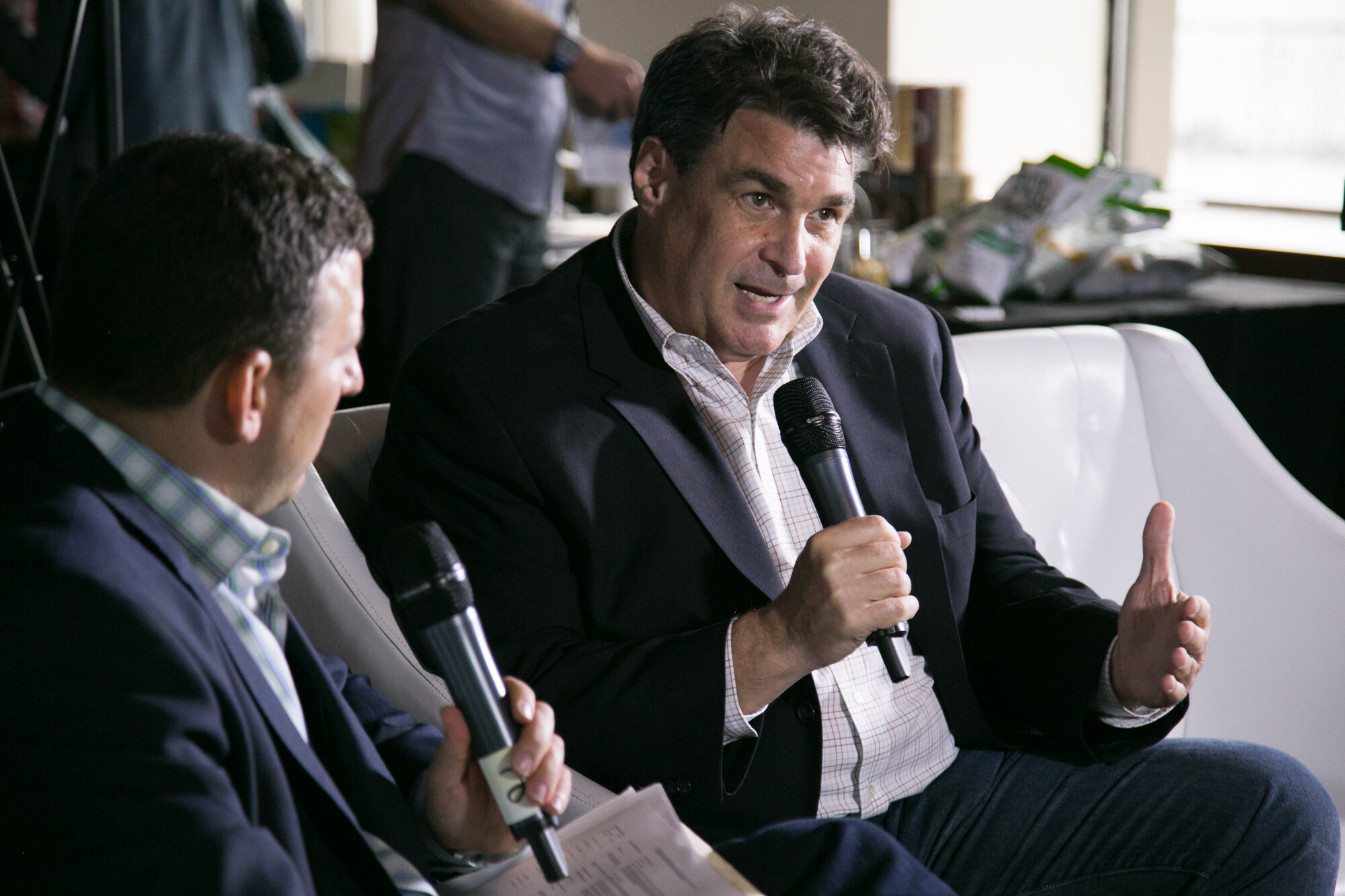 Man in a dark blazer holding a microphone and gesturing, with the shoulder of an interviewer in the foreground and a sample table in the background.