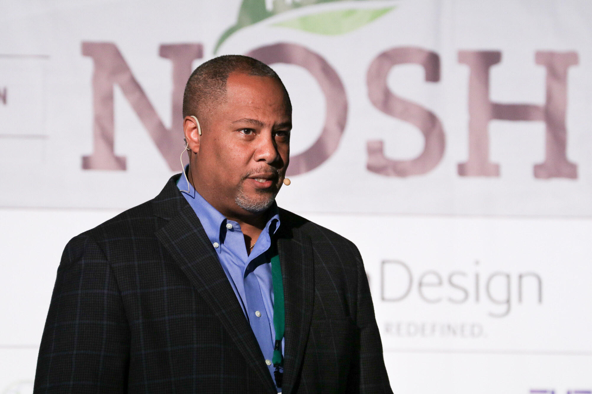 Man in a dark plaid blazer and blue shirt wearing a headset microphone, with part of the Project NOSH logo and SmithDesign text behind him.