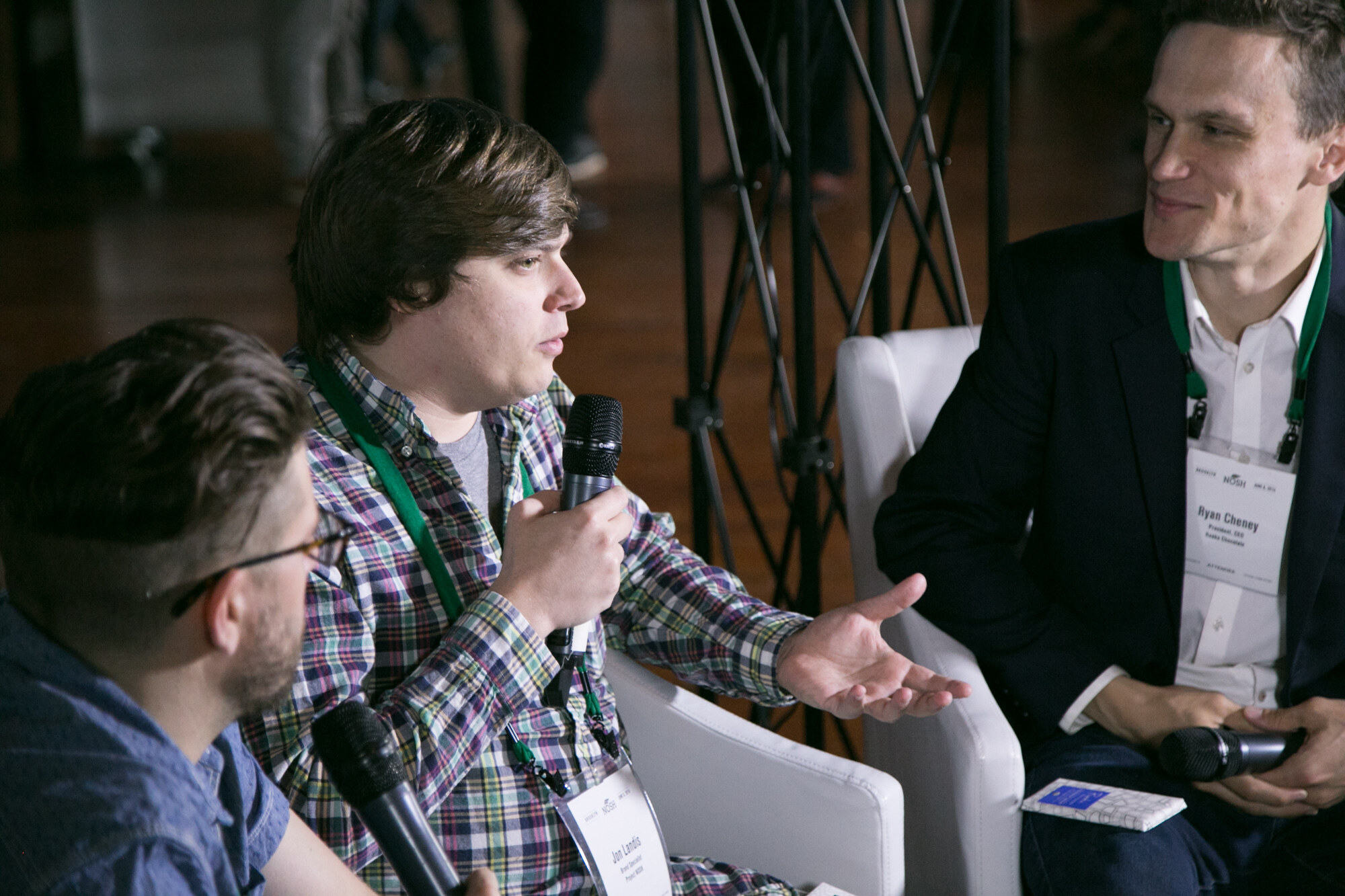 Man in a plaid shirt holding a microphone speaking with another man whose name badge is partly visible, seated in white chairs.