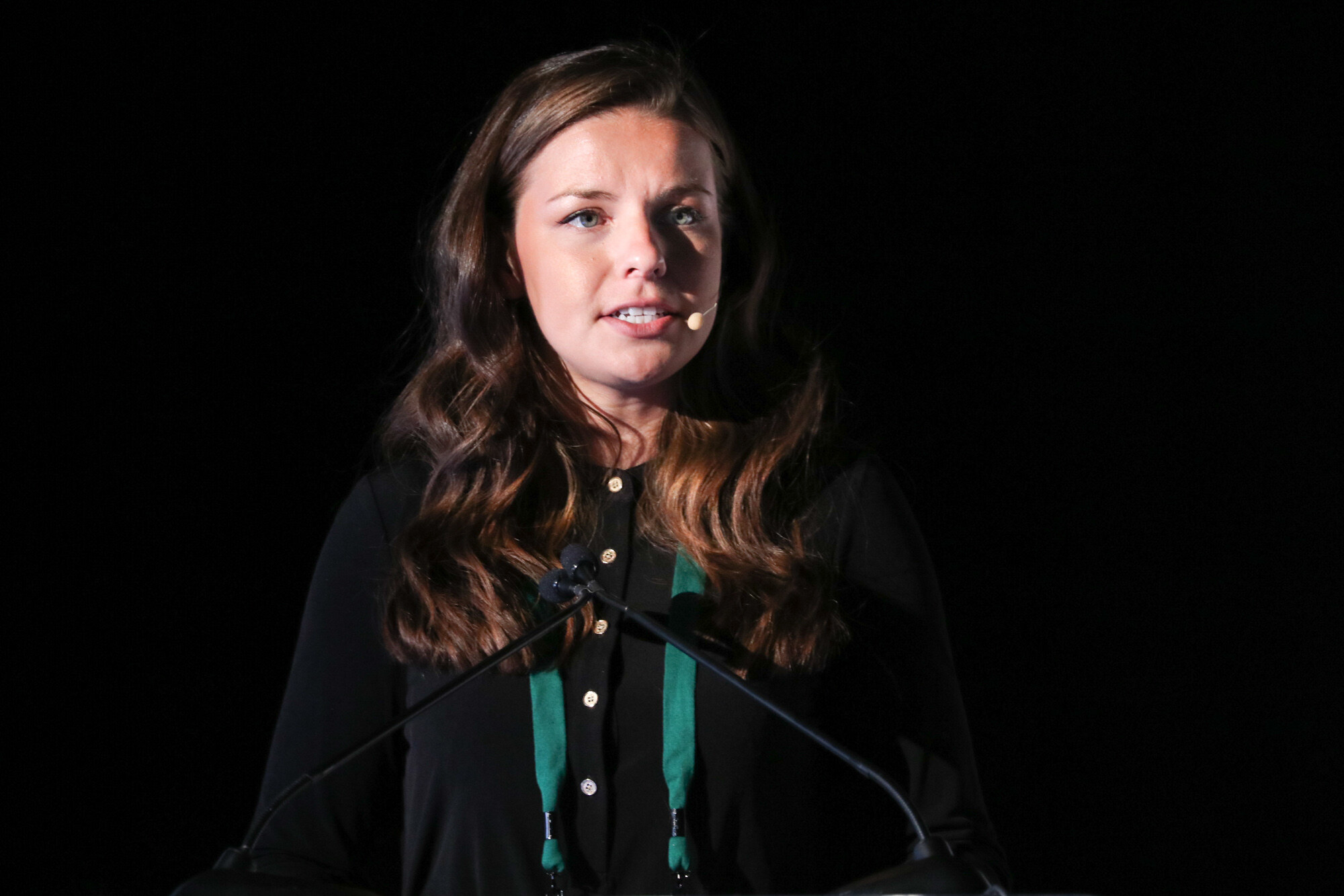 Woman with long wavy brown hair and a headset microphone speaking at a lectern against a black background.