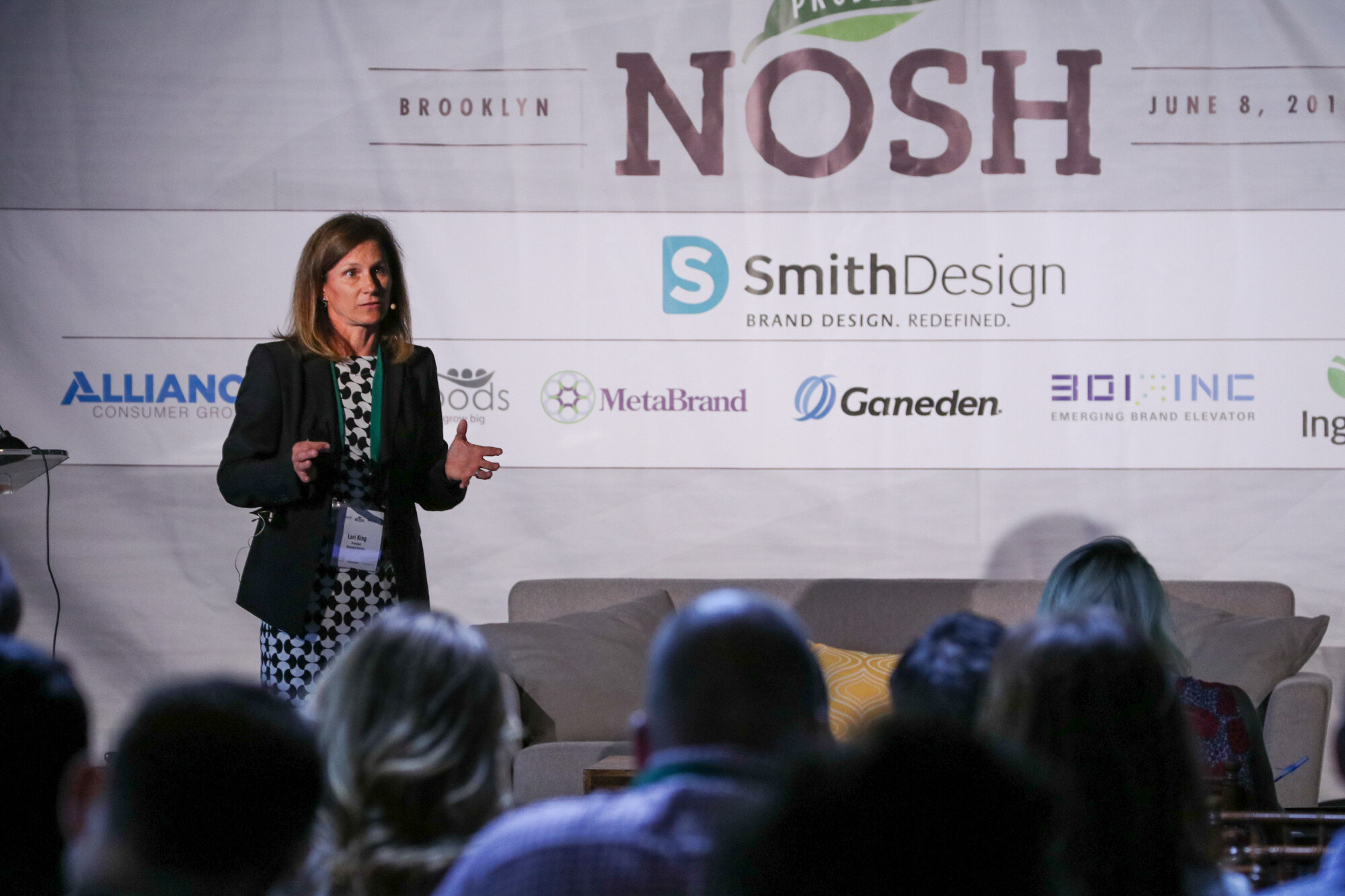Woman in a black blazer and patterned dress speaking beside the stage sofa, with the Project NOSH Brooklyn banner and sponsor logos behind her and audience heads in the foreground.
