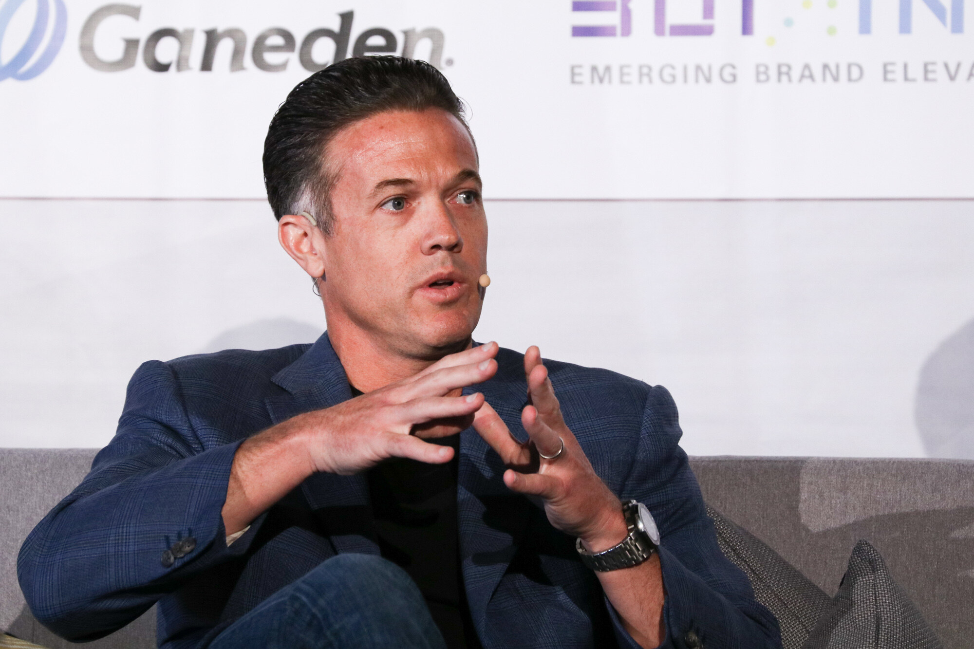 Man in a navy blazer seated on a gray sofa gesturing with both hands, with a Ganeden logo and Emerging Brand Elevator text on the banner behind him.