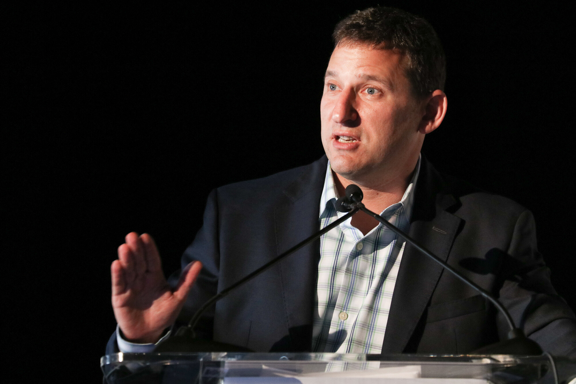 Man in a dark blazer and checked shirt speaking at a clear podium with two microphones against a black backdrop.