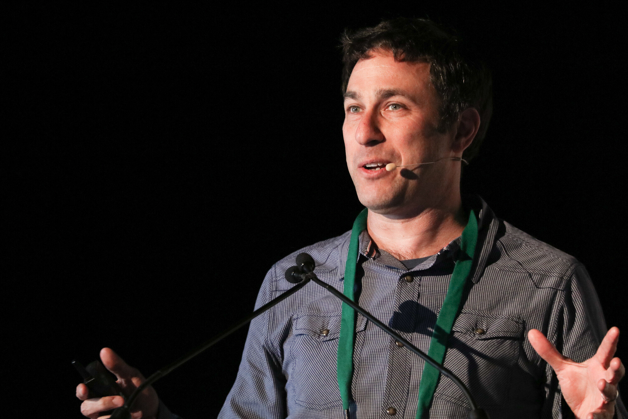 Man in a gray checked shirt and green lanyard speaking at a microphone with one hand open and a slide clicker in the other.