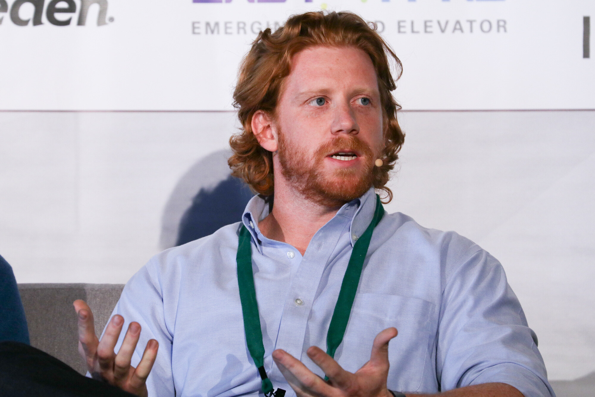 Man with red hair and a beard in a light blue shirt and green lanyard gesturing while speaking, with Emerging Brand Elevator text on the banner behind him.