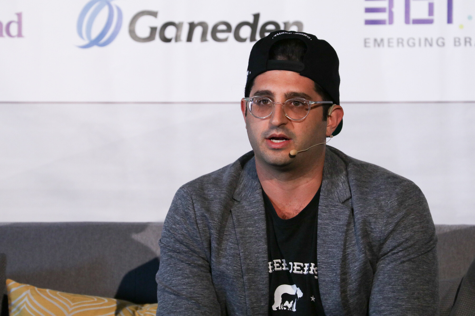 Man in a backwards black cap, clear-framed glasses and a gray jacket speaking into a headset microphone, with a Ganeden logo behind him.