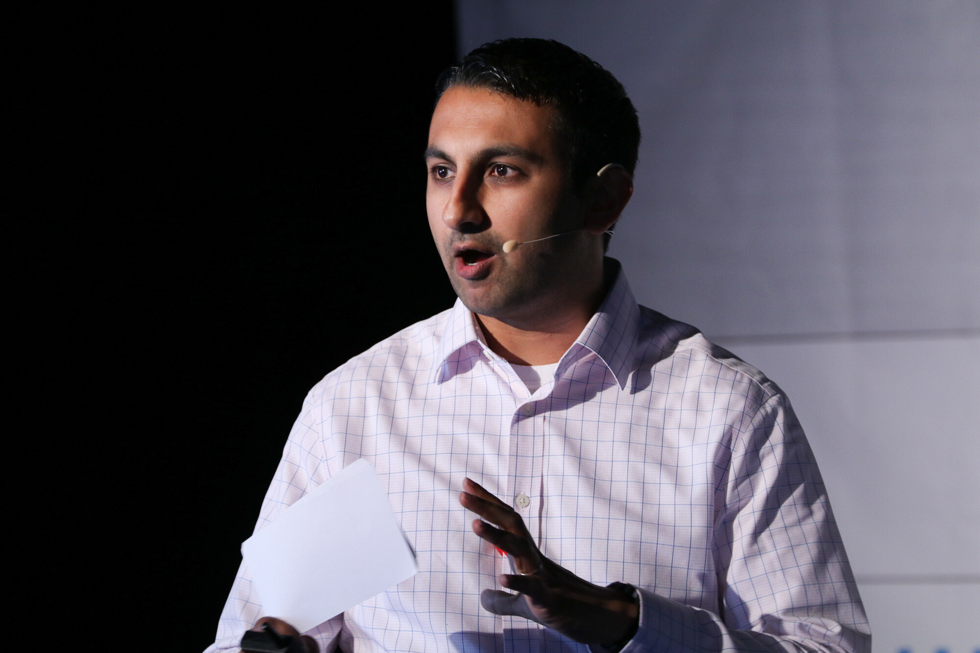 Man in a light checked shirt holding paper in one hand and gesturing with the other while speaking.