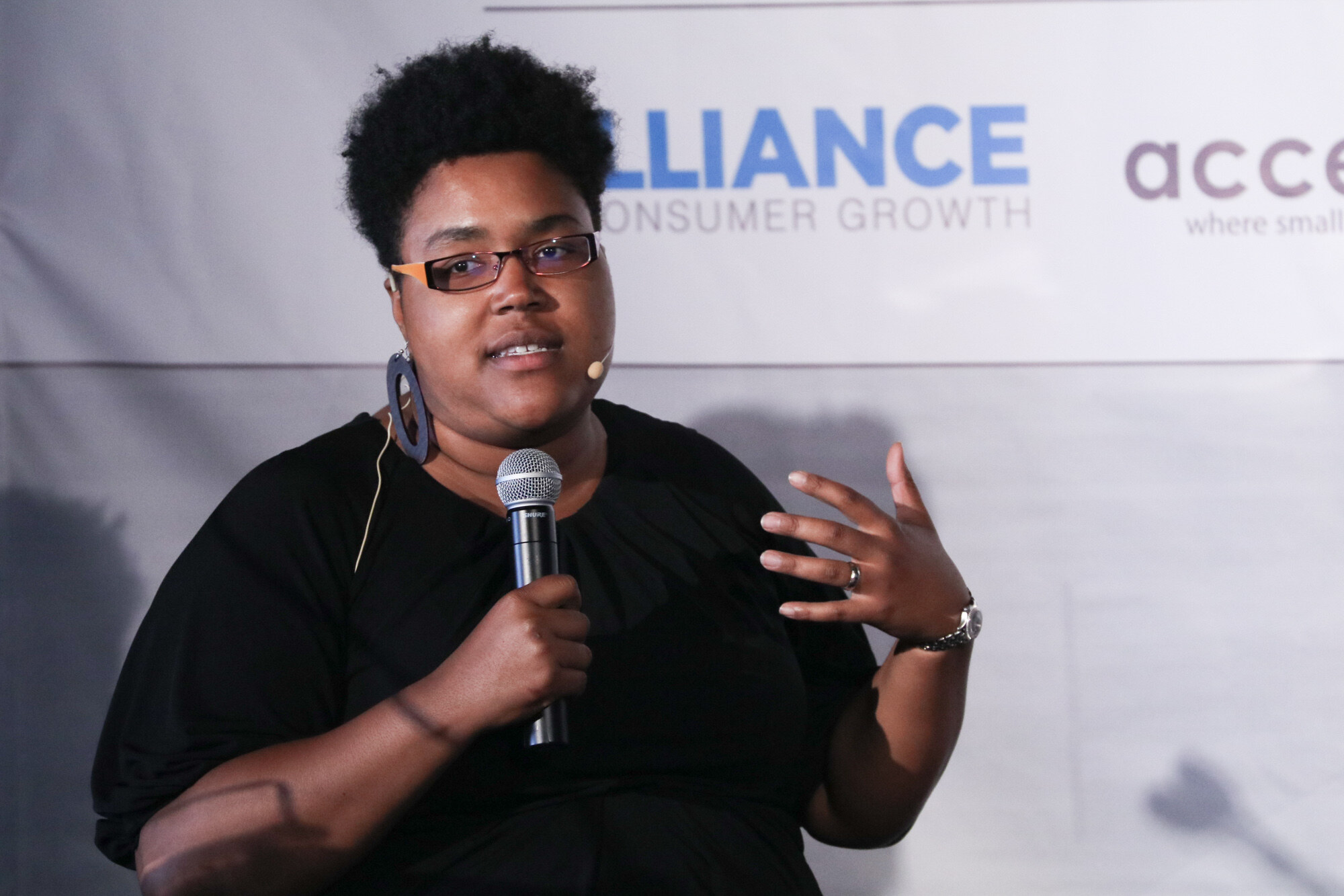 Woman with short natural hair and glasses holding a handheld microphone and gesturing, with Alliance for Consumer Growth and accelFoods text on the banner behind her.