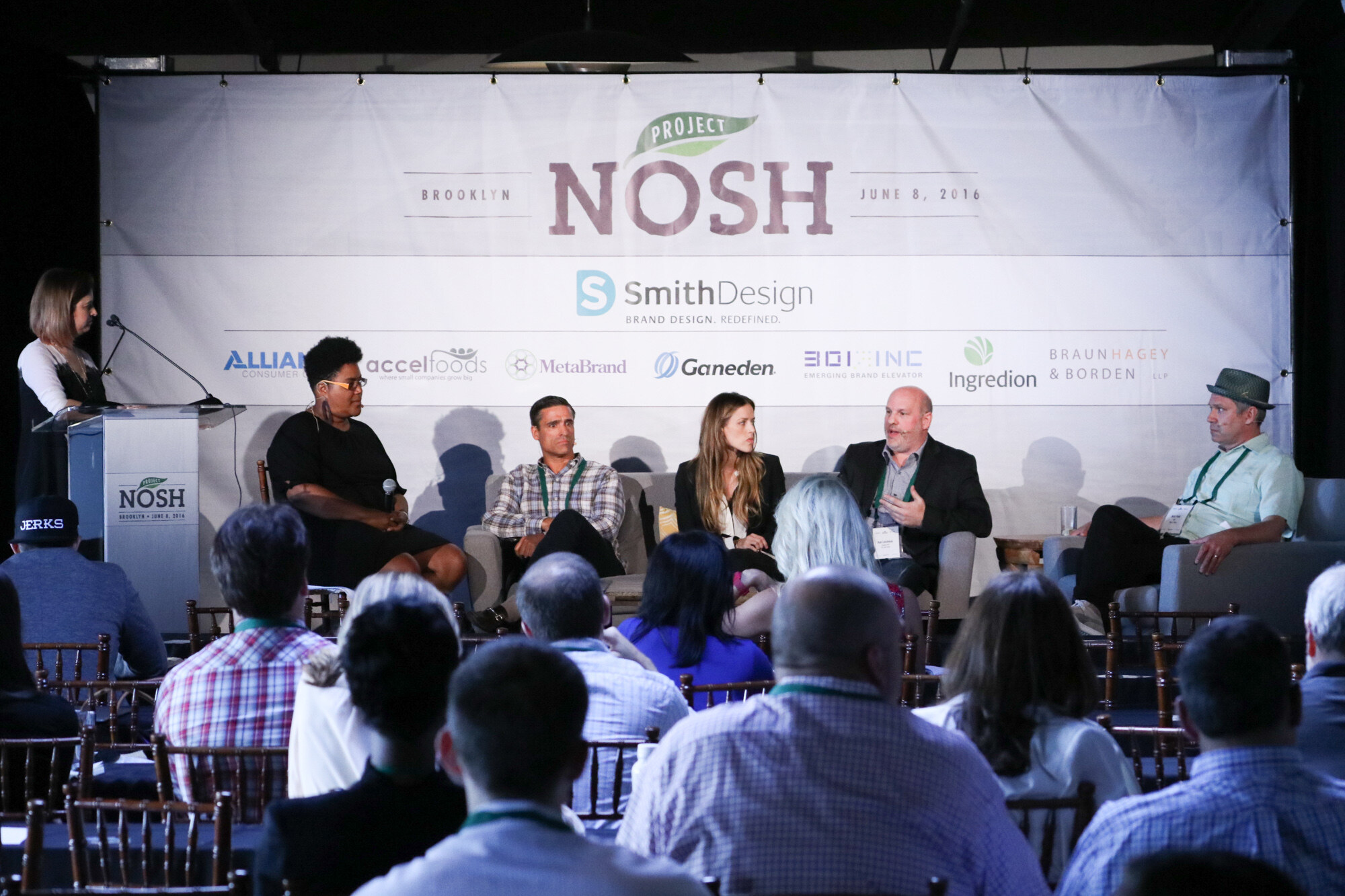 Wide view of five panelists seated on stage sofas under the Project NOSH Brooklyn banner, with a woman at a podium on the left and rows of seated attendees in the foreground.