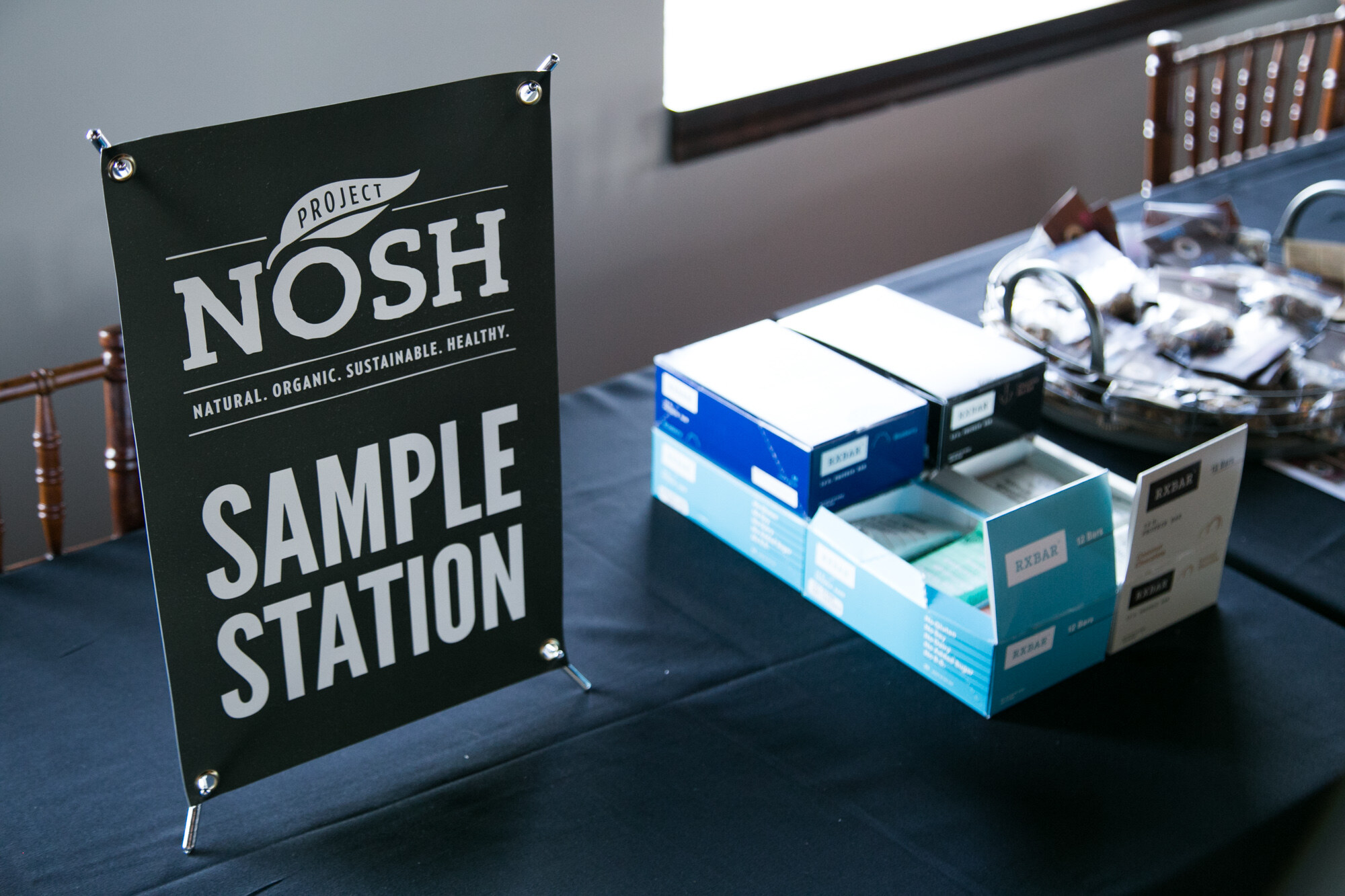 Black Project NOSH Sample Station sign on a table with open blue boxes of RXBAR protein bars beside it.