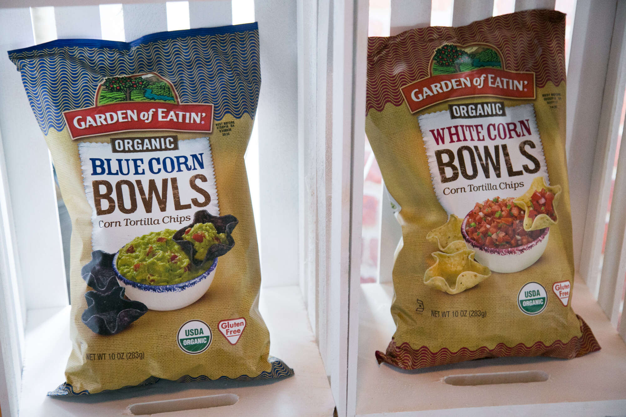 Two bags of Garden of Eatin Organic tortilla chip bowls, one blue corn and one white corn, in a white crate.