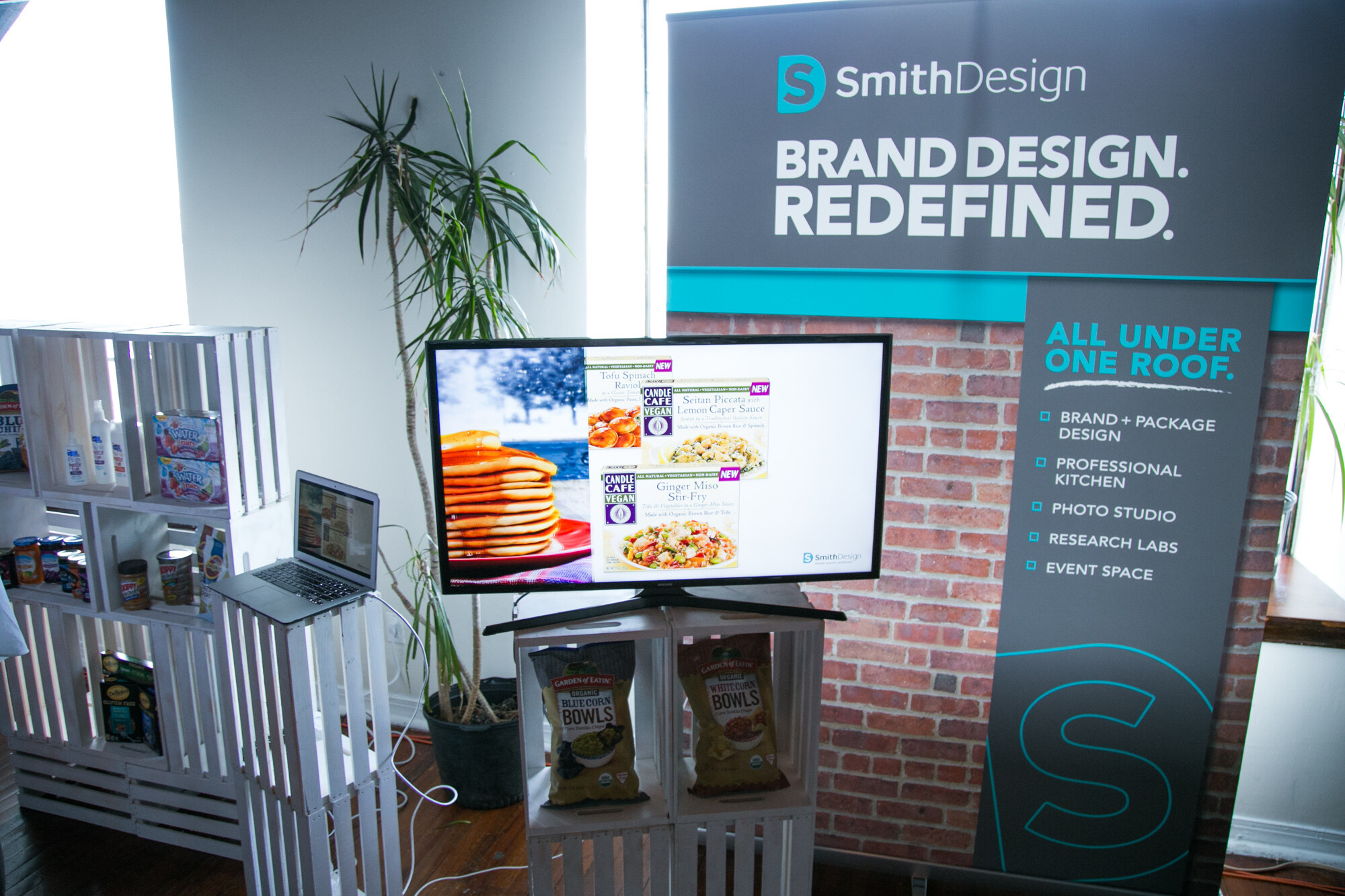 Exhibit space with a SmithDesign Brand Design Redefined banner, a TV showing packaged food images, a laptop and white crate shelving with Garden of Eatin bags.