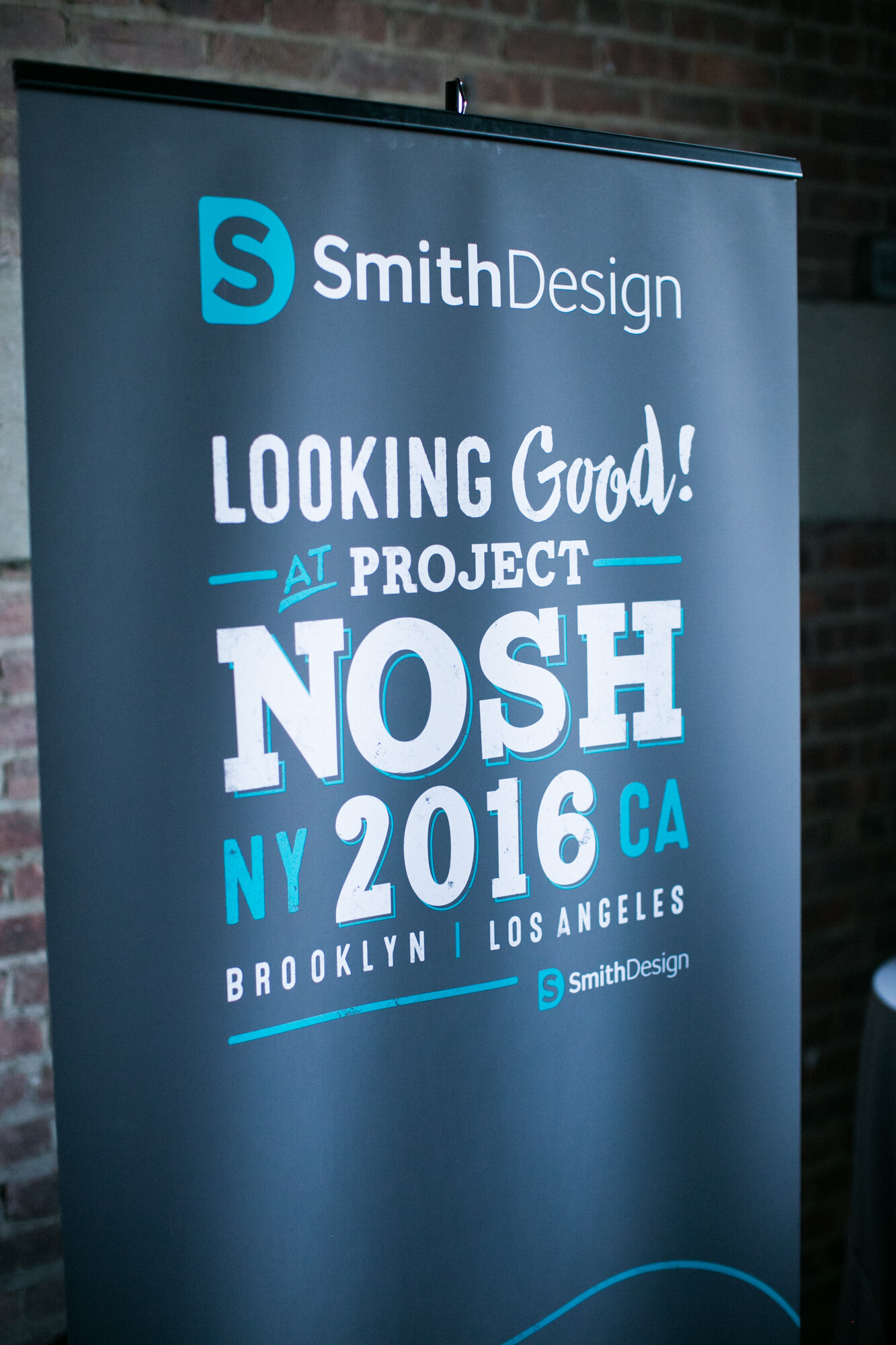 Dark gray pull-up banner reading SmithDesign, Looking Good at Project NOSH NY 2016 CA, Brooklyn and Los Angeles, against a brick wall.