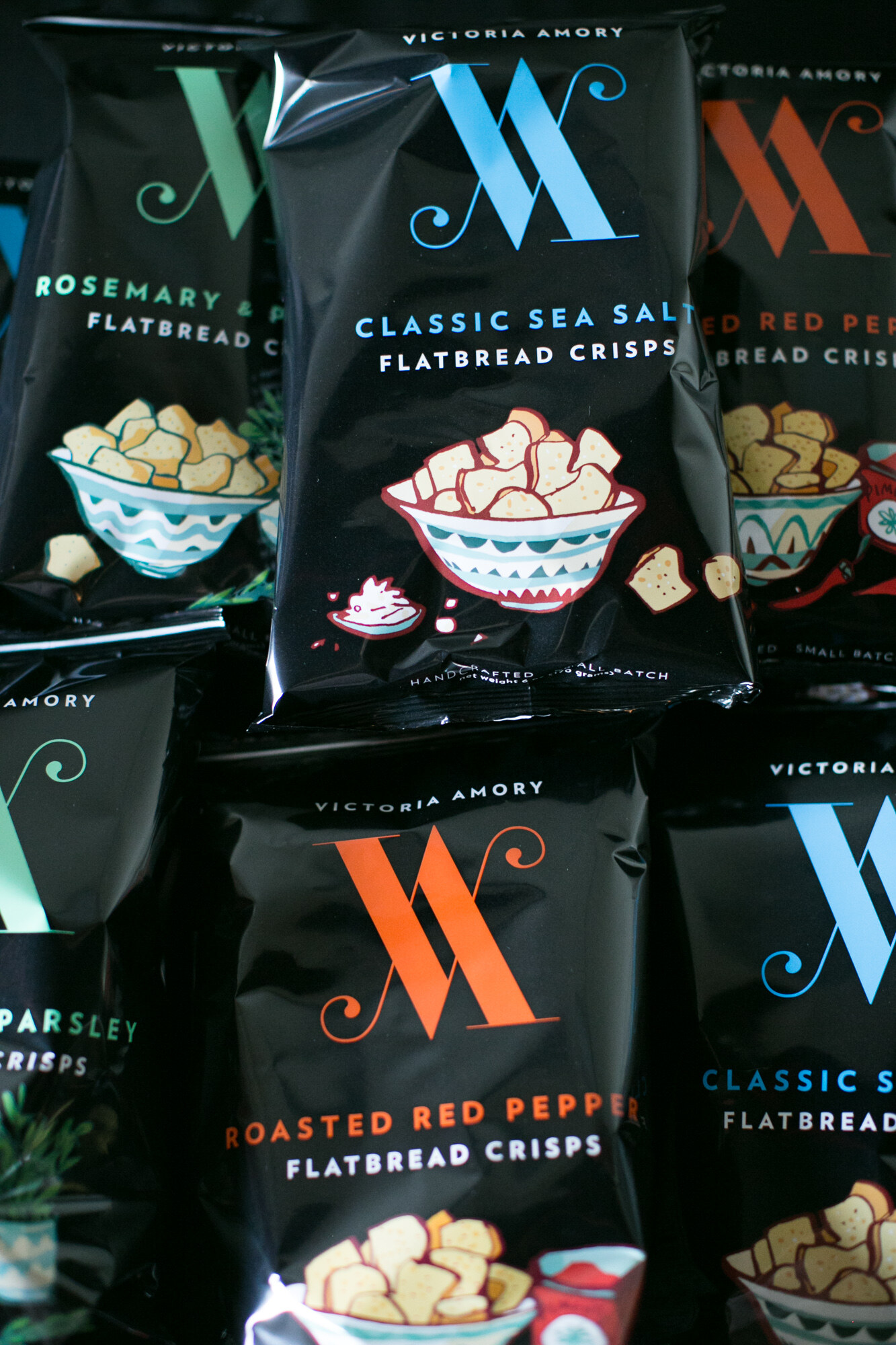 Stacked black bags of Victoria Amory Flatbread Crisps in Classic Sea Salt and Roasted Red Pepper flavors.