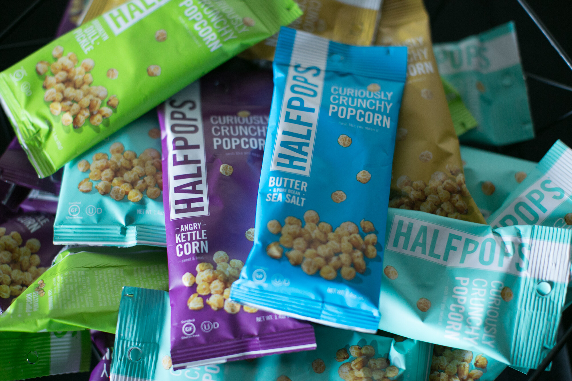 Pile of green, teal, purple and blue Halfpops popcorn packs including Angry Kettle Corn and Butter and Sea Salt.