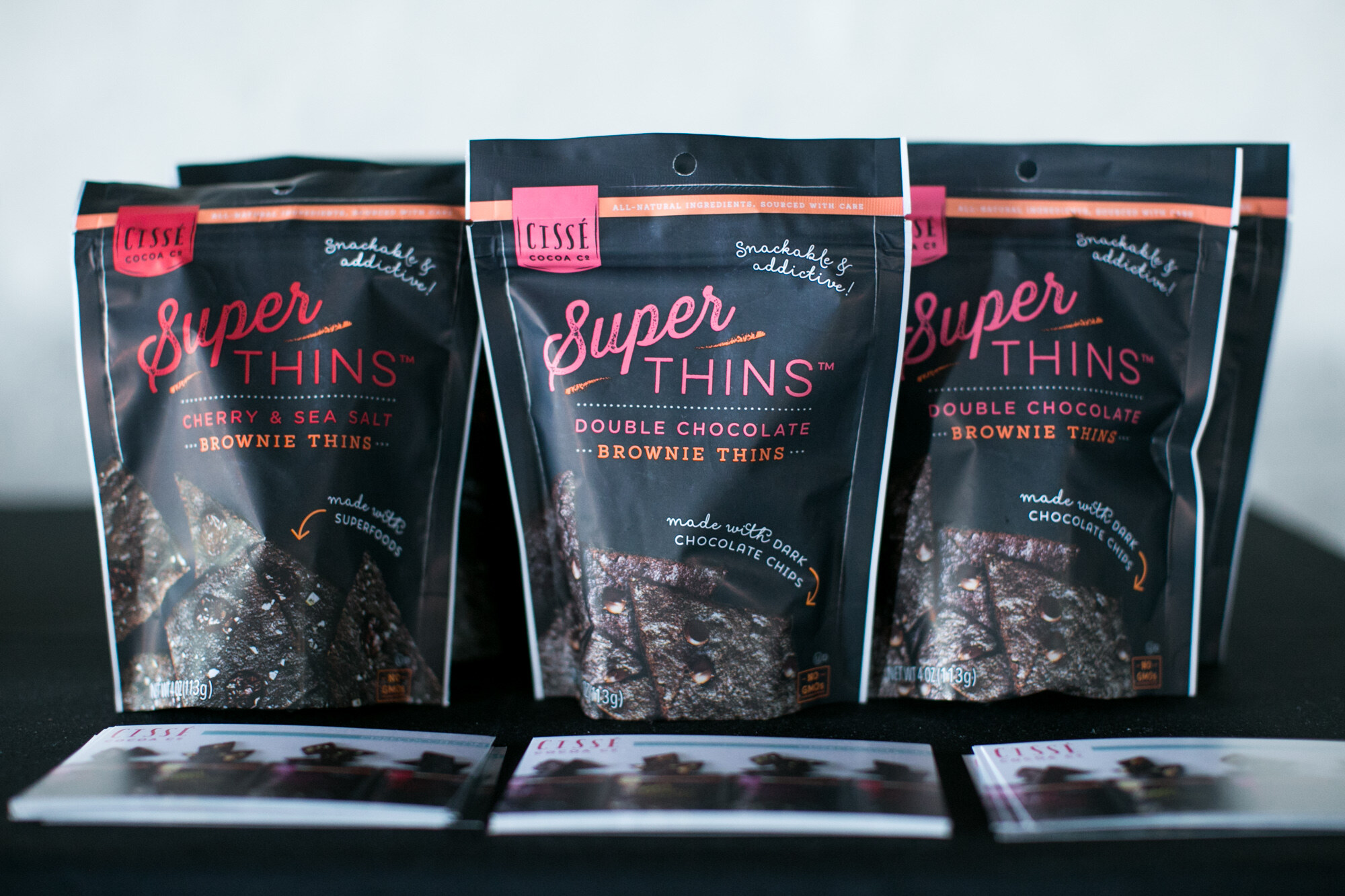 Three black bags of Cisse Cocoa Co. Super Thins in Cherry and Sea Salt and Double Chocolate flavors with printed cards below.