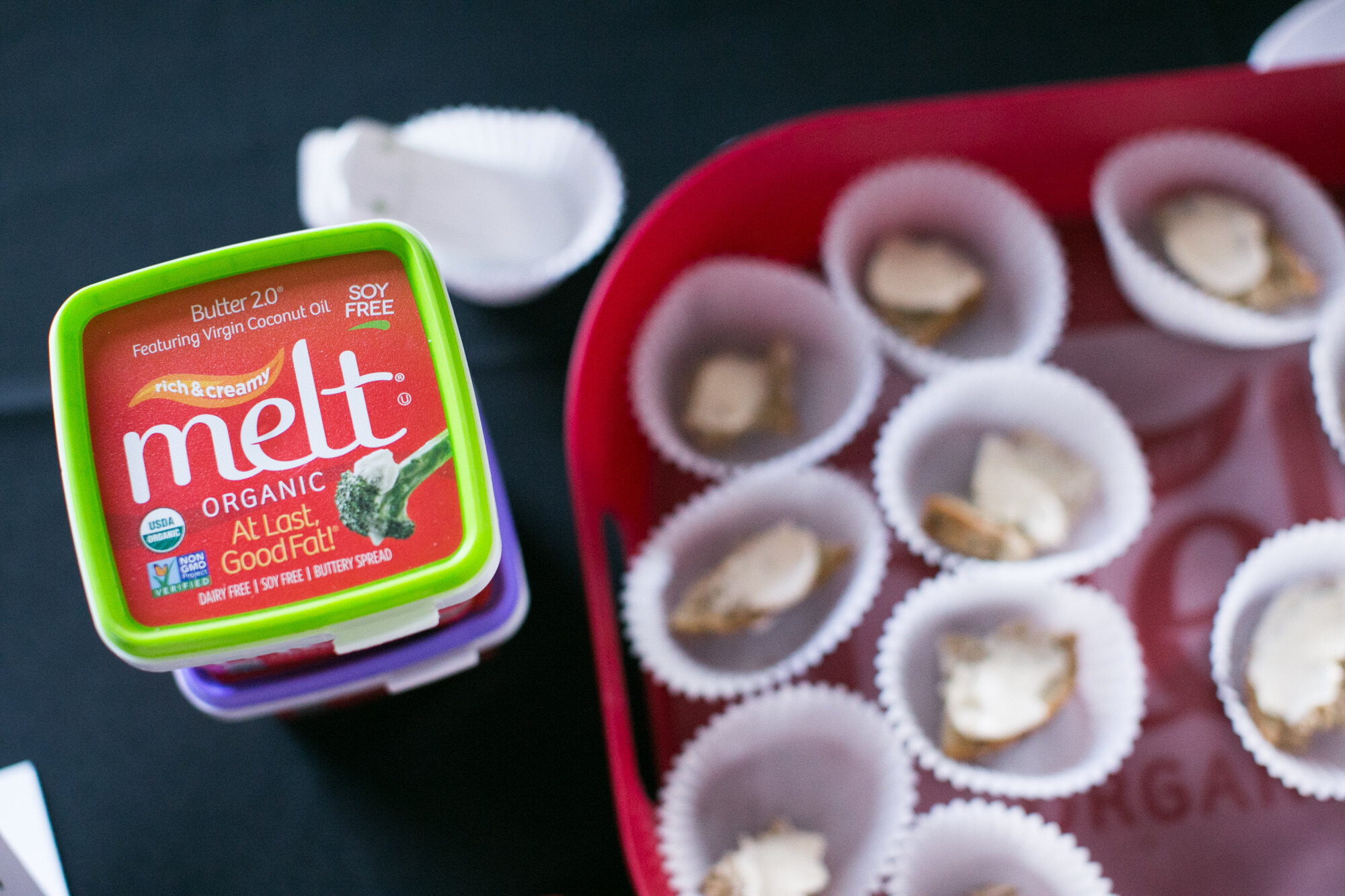 Overhead view of a green-lidded Melt Organic Butter 2.0 tub beside a red tray of paper cups holding small spread samples.