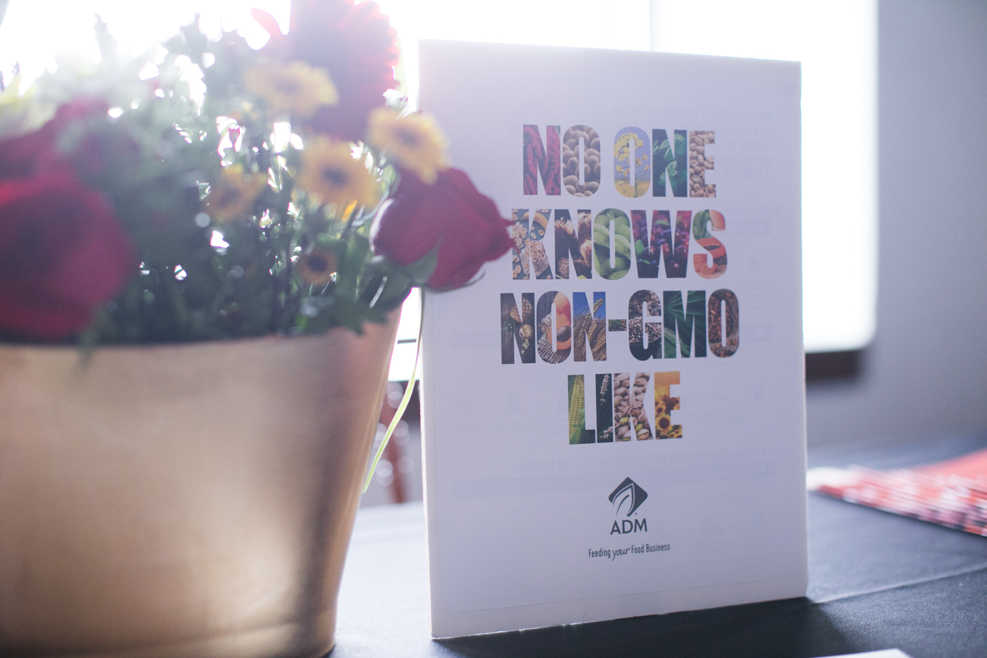 Printed card with the text No One Knows Non-GMO Like and the ADM logo next to a pot of red and yellow flowers.