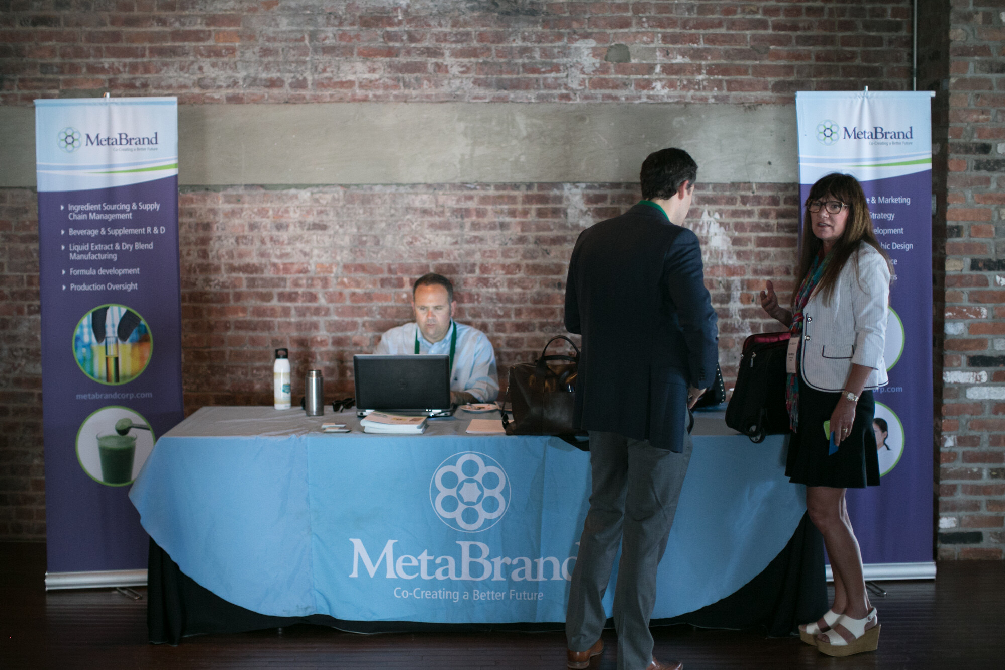 MetaBrand table with a light blue tablecloth and two vertical MetaBrand banners, a seated man at a laptop, and two people talking in front.