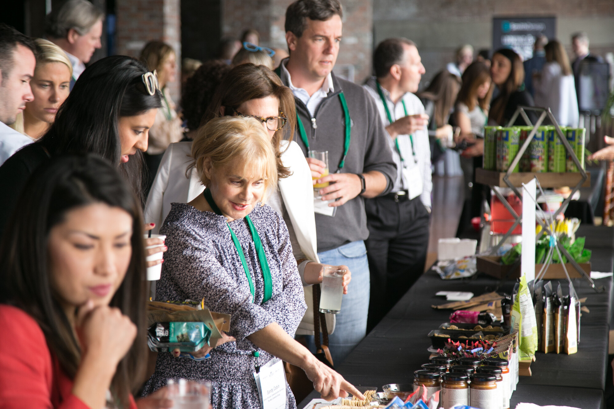 Woman in a patterned dress pointing at items on the sample table while other attendees look on.