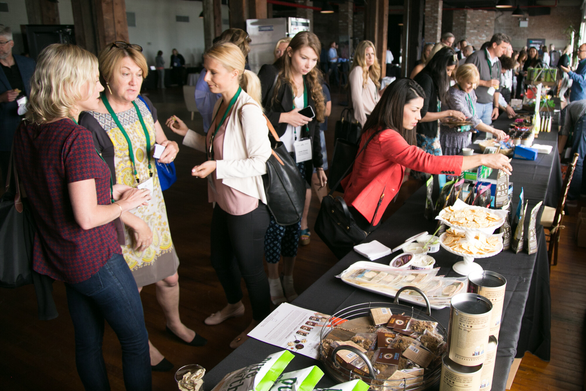 Group of attendees wearing green lanyards browsing samples on a long black-draped table.