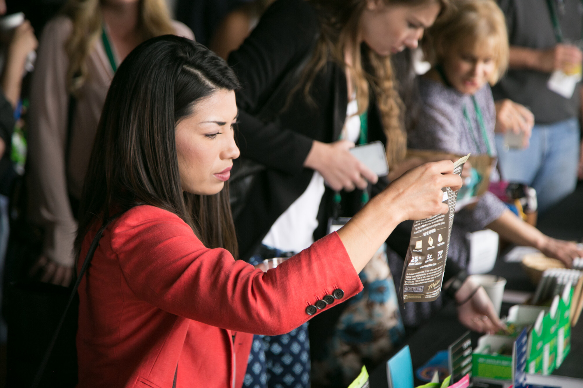 Woman in a red blazer lifting a package to read it at a crowded sample table.
