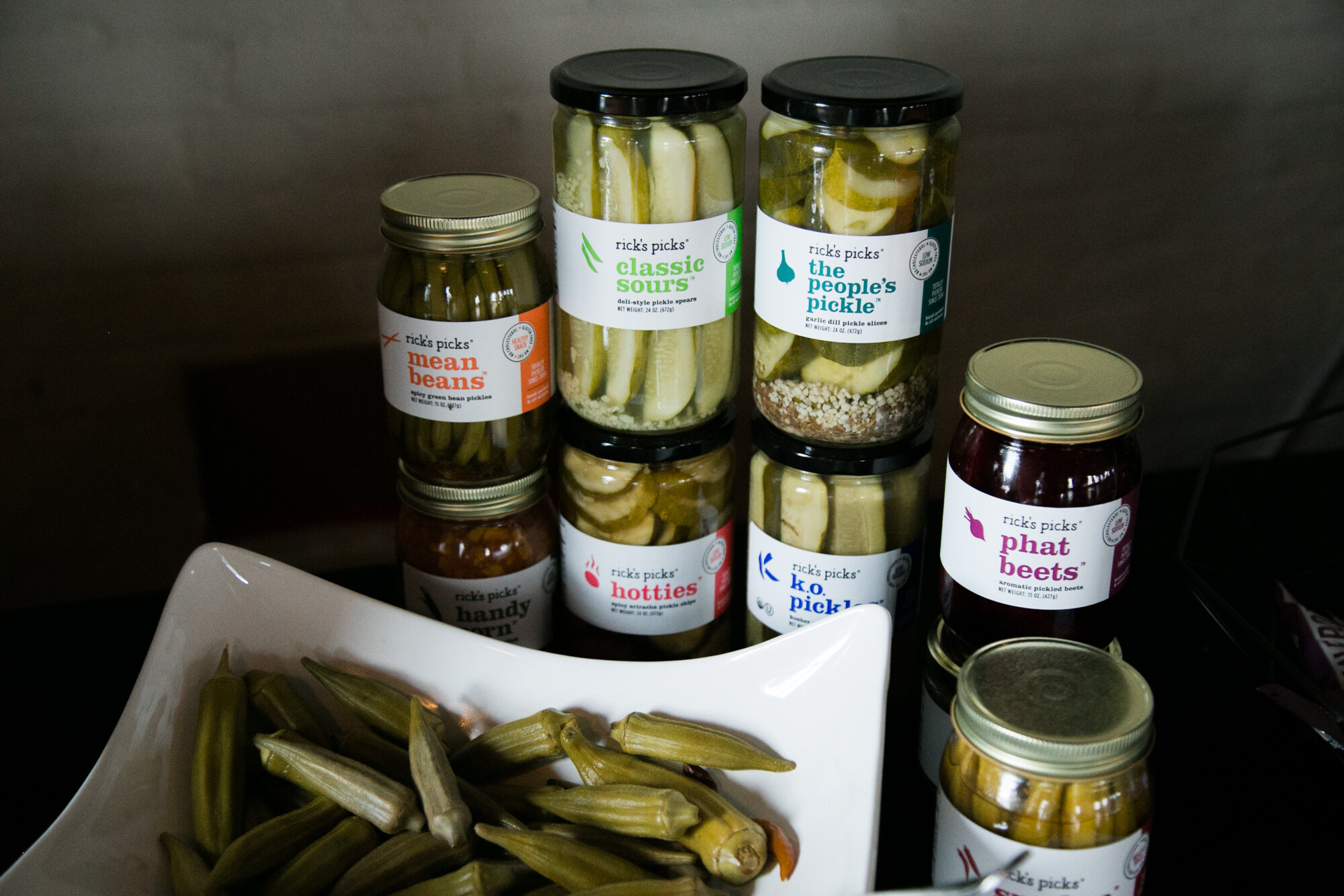 Jars labeled Rick's Picks Mean Beans, Classic Sours, The People's Pickle, Hotties and Phat Beets, with pickled okra in a white dish.