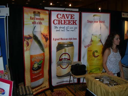 A booth with tall banners for Cave Creek Chili Beer, Dorada beer and a lemonade drink. A woman with dark curly hair stands beside a table, and a black bag sits on a chair.