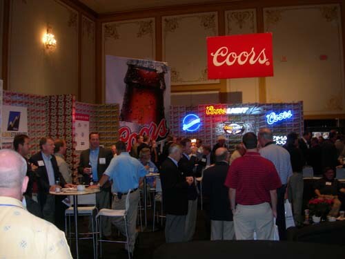 Attendees stand and talk in a hall near a red Coors sign, a large bottle graphic and lit neon signs for Coors Light and other beer brands. Some stand at tall tables.