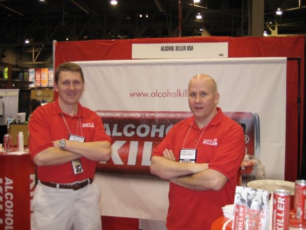 Two men in red Alcohol Killer polo shirts stand with arms crossed in front of a banner at the Alcohol Killer USA booth, with Alcohol Killer cans on a table nearby.
