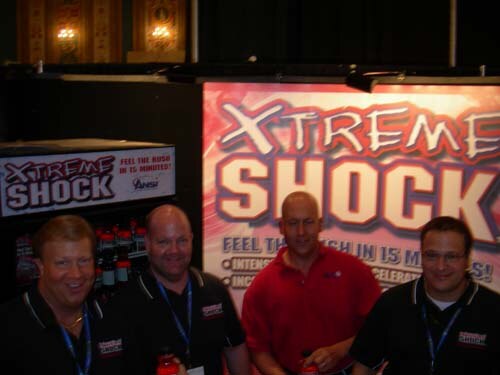 Four men pose together at the Xtreme Shock booth. Three wear black shirts with lanyards, and one wears a red shirt. A large Xtreme Shock banner is behind them.