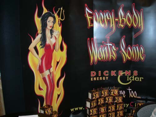 A dark booth backdrop shows an illustrated woman in a red dress in front of flames. Text reads "Every-body Wants Some" and Dickens Energy Cider, with boxes stacked below.