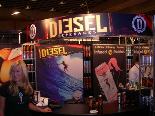 A booth with a Diesel Beverages sign overhead and Diesel Energy Drink and 3 SUM banners. A blonde woman in a black shirt stands in front, and cans sit on shelves and the counter.
