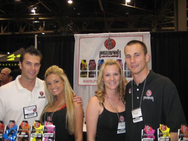Two men and two women in a row smiling at a Powerock booth, wearing badges on lanyards, with a Powerock banner behind them and rows of Powerock energy shot bottles in front.