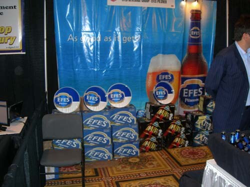 A booth with a blue Efes Pilsener banner showing a bottle and glass of beer. Cases of Efes are stacked on the floor, and a man in a dark suit stands at right.